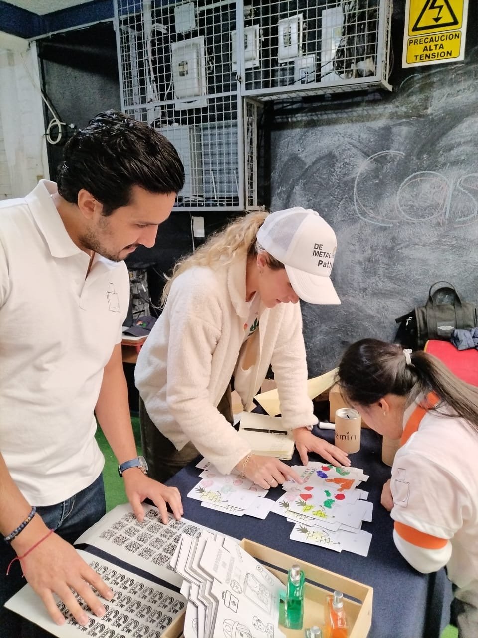 Three people in casual attire sorting colorful sheets and stickers on a table, with a caution sign in the background.