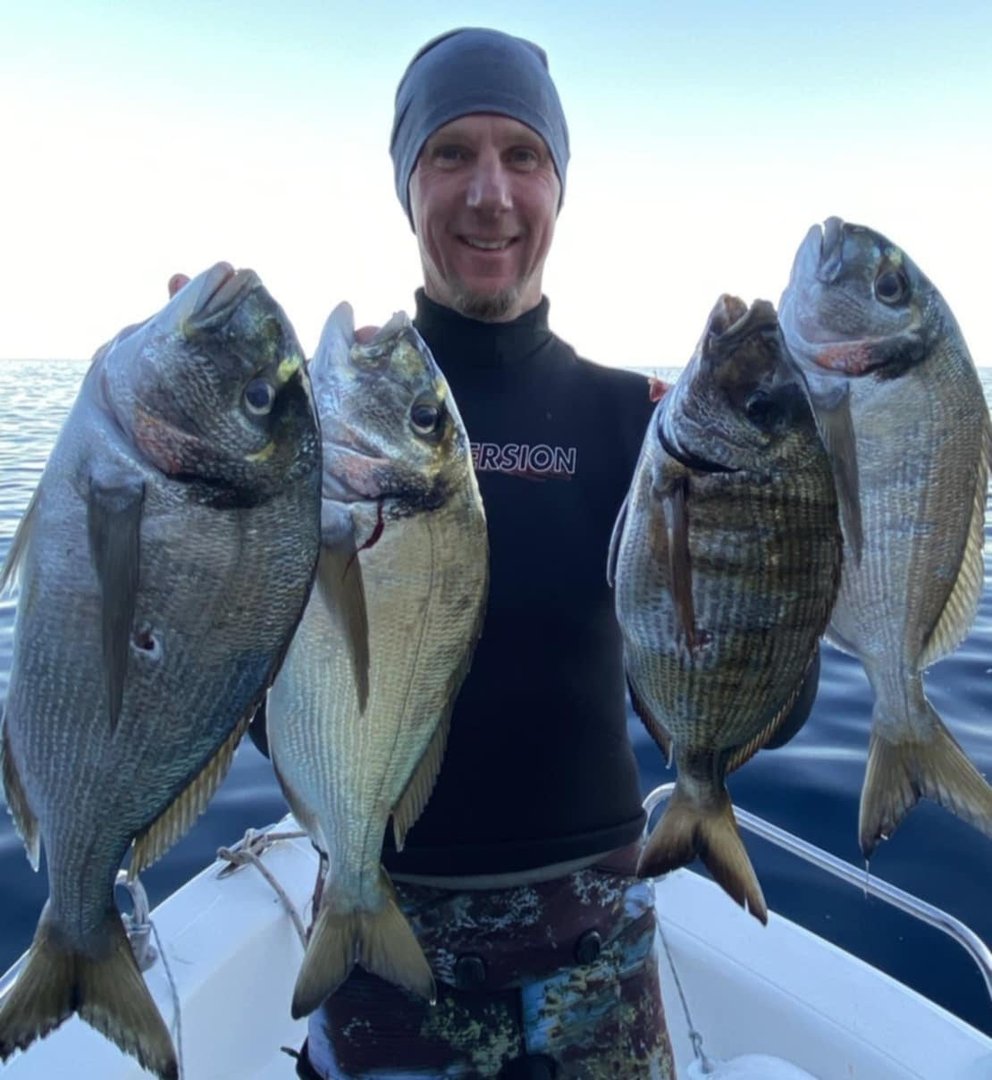 Person on a boat holding four large fish, wearing a wetsuit and beanie, with a calm ocean in the background.
