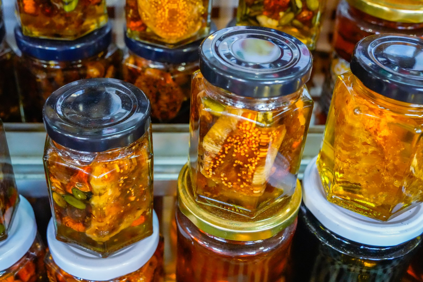 Close-up view of honey jars with walnuts, almonds, fruit slices, and honeycomb in golden honey at a rustic food market.