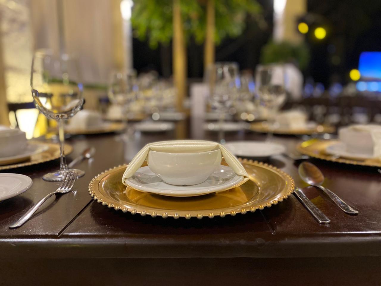 Elegantly set dining table with gold-rimmed plates, folded napkins, glasses, and silverware in a dimly lit, classy setting.
