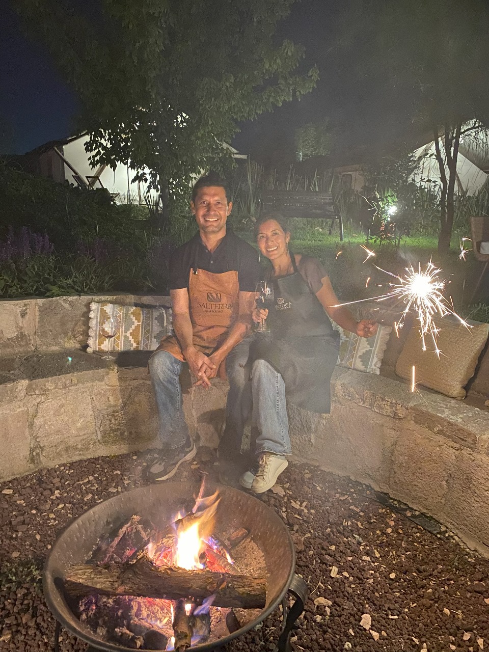 Two people sitting by a fire pit at night, one holding a sparkler, smiling, surrounded by greenery and dimly lit tents.