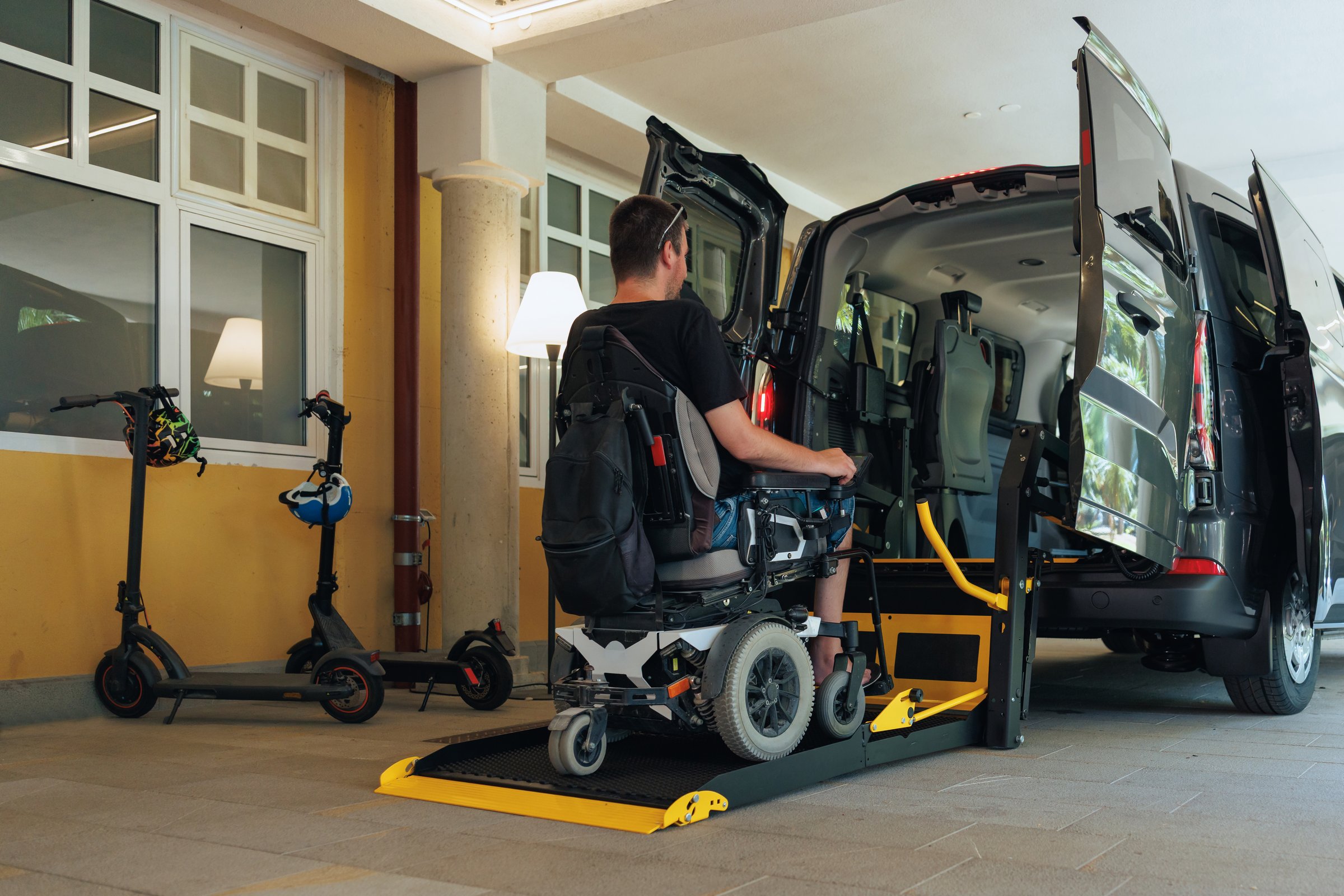 Person with disability accesses a van using a wheelchair lift in a parking area. The setting is urban and designed for inclusivity.