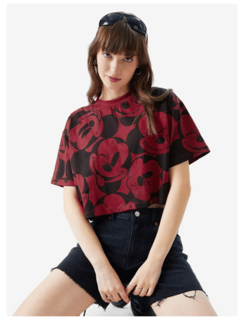 Woman in a red Mickey Mouse print t-shirt and denim shorts, wearing sunglasses on her head, poses confidently.