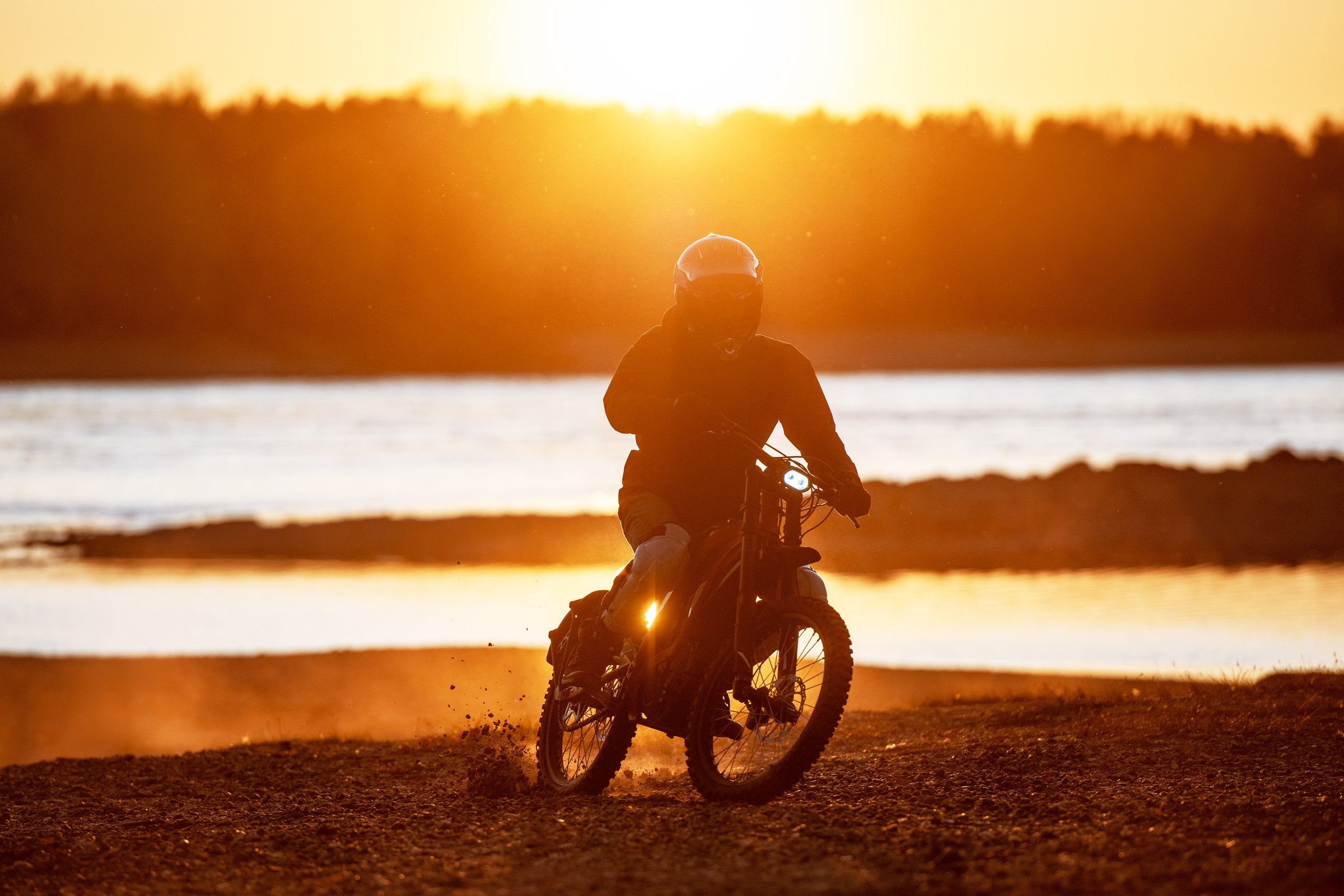 Motorcyclist rides on electric enduro motorbike against river and sunset light