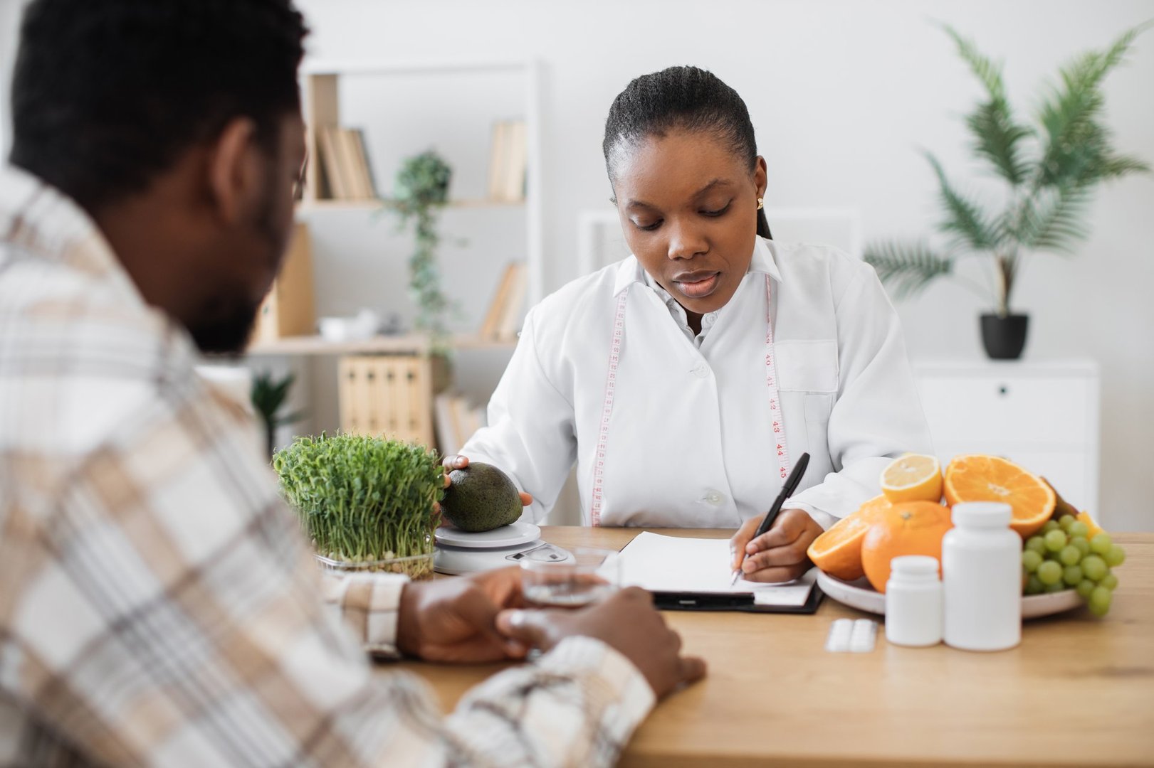 Focused female specialist writing details about fruit nutrients on clipboard paper while counselling client. Multicultural expert in diet formulating special meals for male patient in clinic.