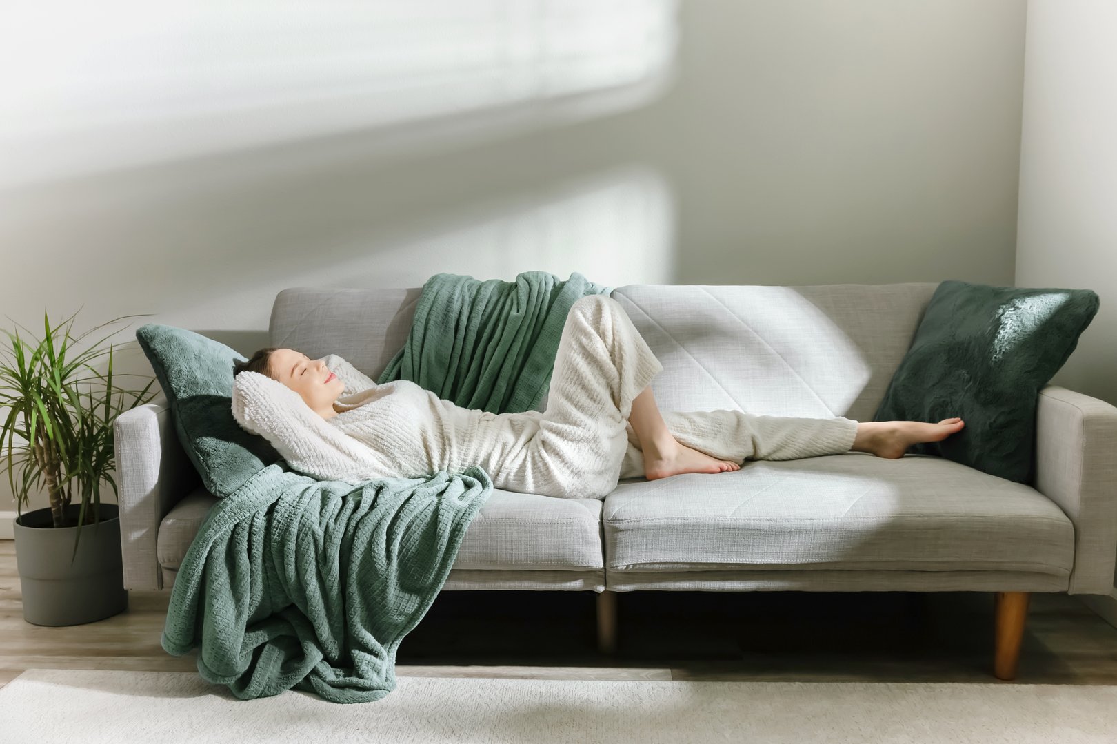 Woman in soft loungewear resting on a modern sofa with green blankets and pillows. Natural light and shadows create a peaceful, cozy mood. Ideal for wellness, lifestyle, or home interior visuals.