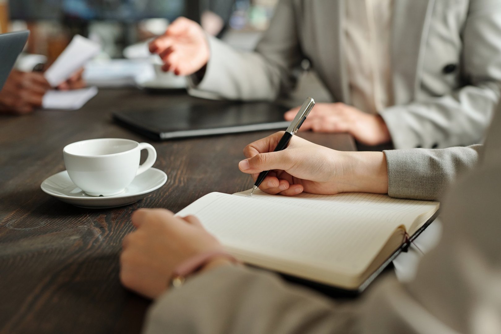 Caucasian young adult woman writing in notebook during business meeting, sitting at table with coffee cup, another business professional gesturing in background, top view