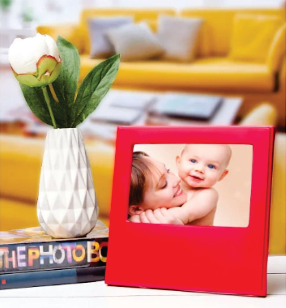 A framed photo of a woman with a baby beside a white vase with a rose, placed on a stack of books in a room with a yellow sofa.