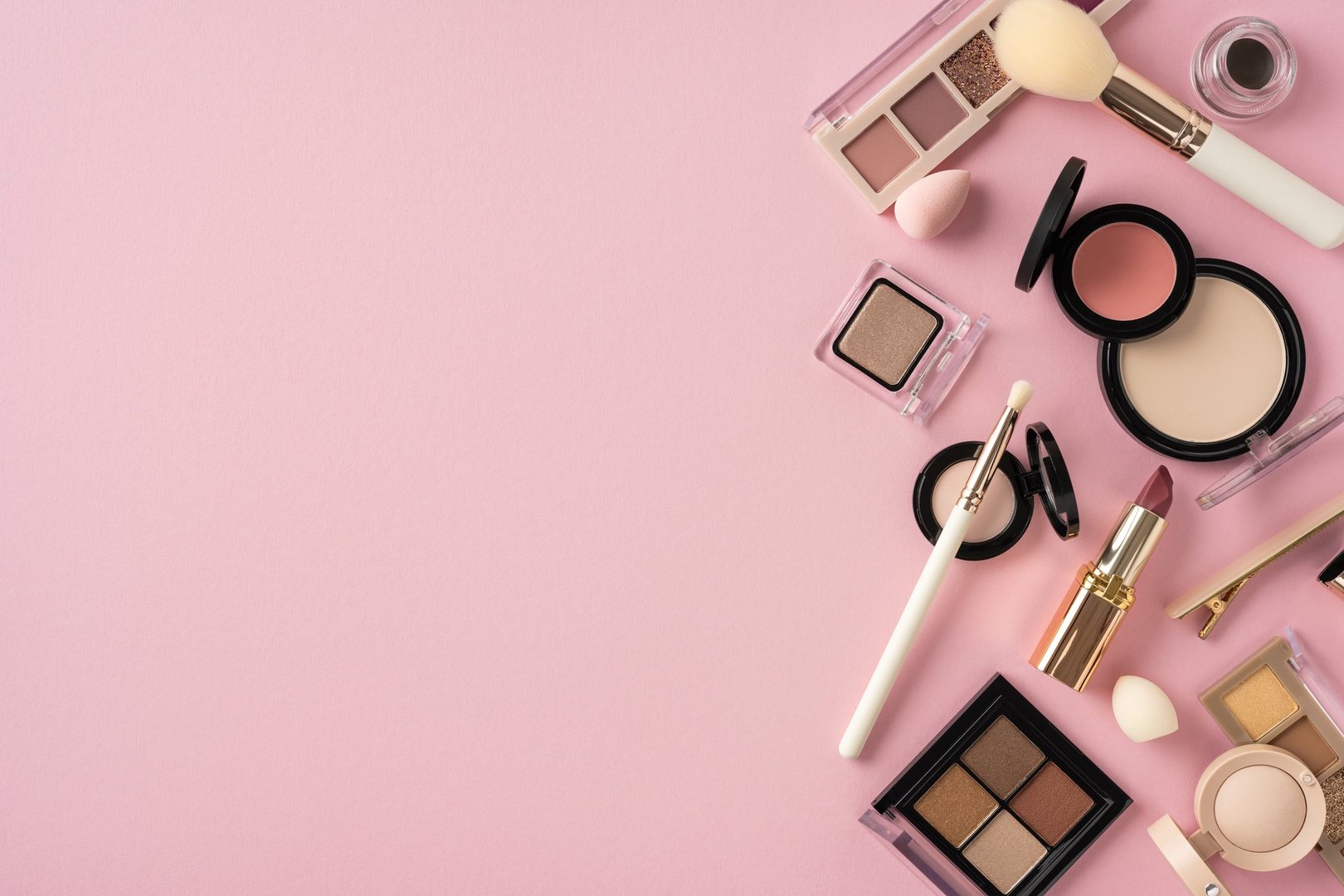 Assorted cosmetic items, including brushes, eyeshadows, and lipsticks, laid on pink backdrop