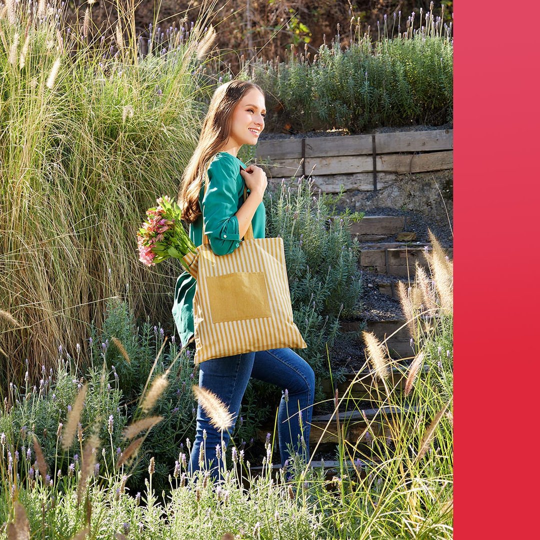 Woman walking outdoors with a yellow tote bag and flowers, surrounded by green plants and wooden steps.
