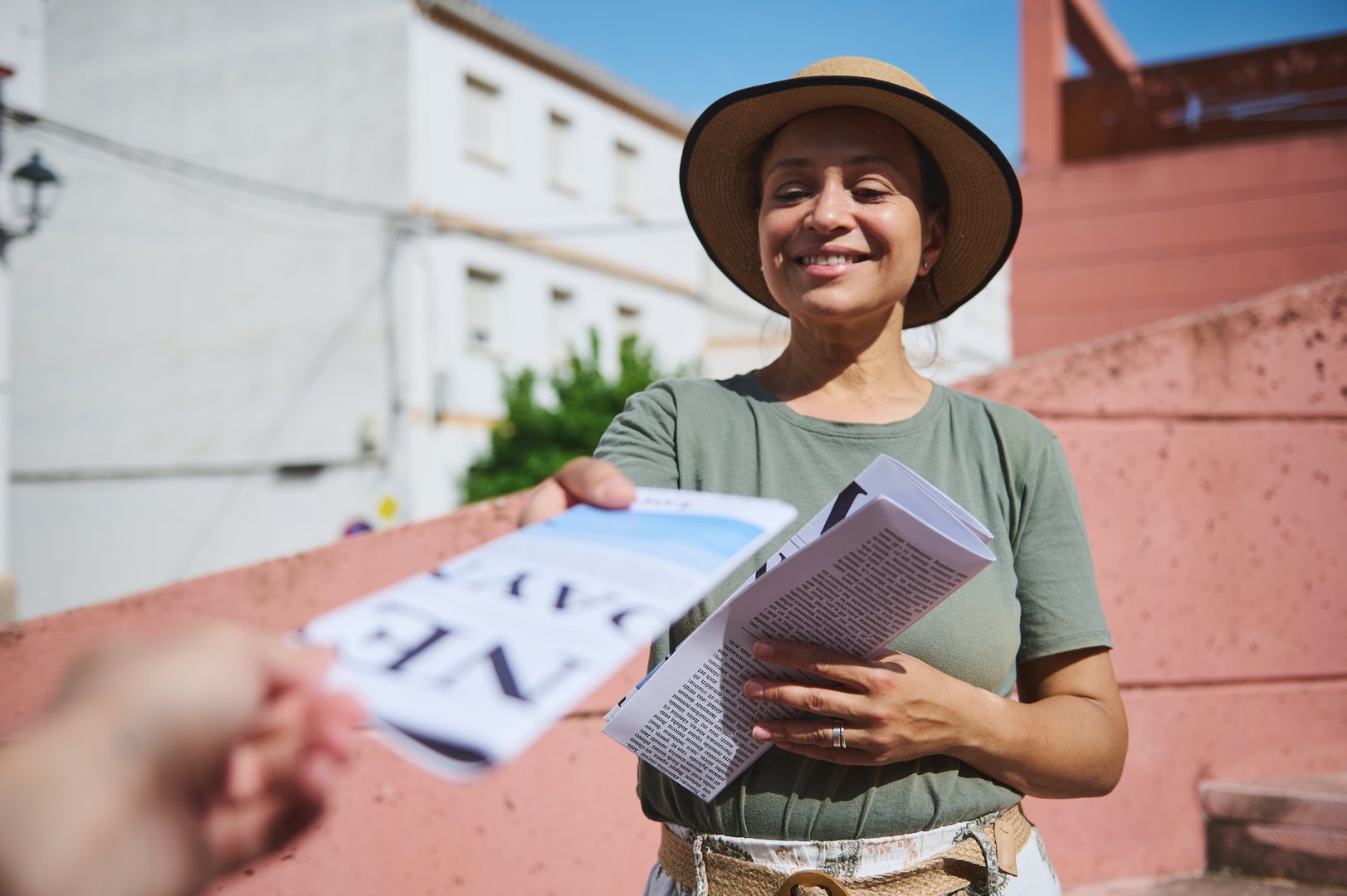A cheerful woman wearing a hat and green shirt hands out flyers outdoors in a sunny urban setting. The image conveys friendliness, engagement, and community interaction.