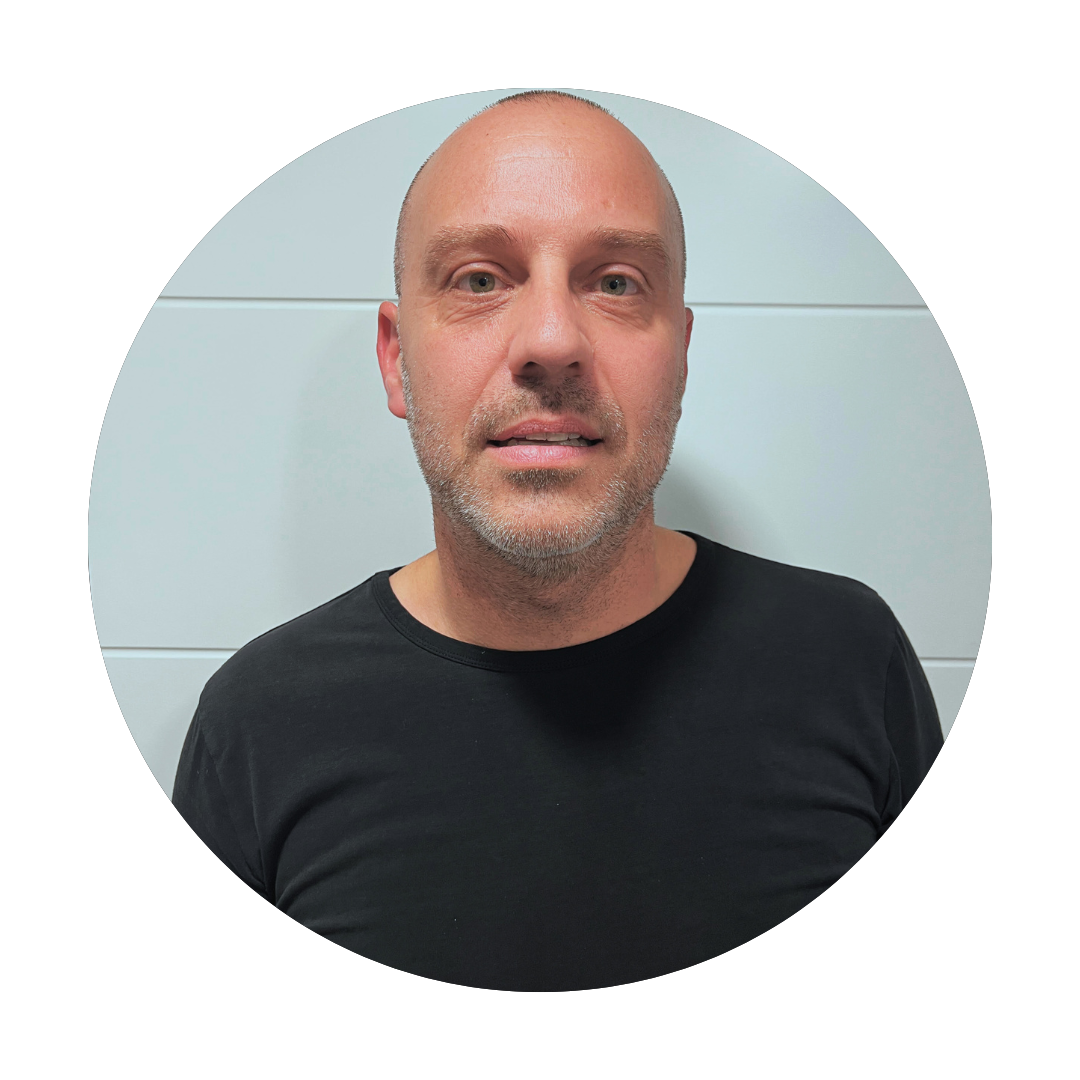 A man with a shaved head and stubble wearing a black shirt, standing against a light tiled wall, looking directly at the camera.