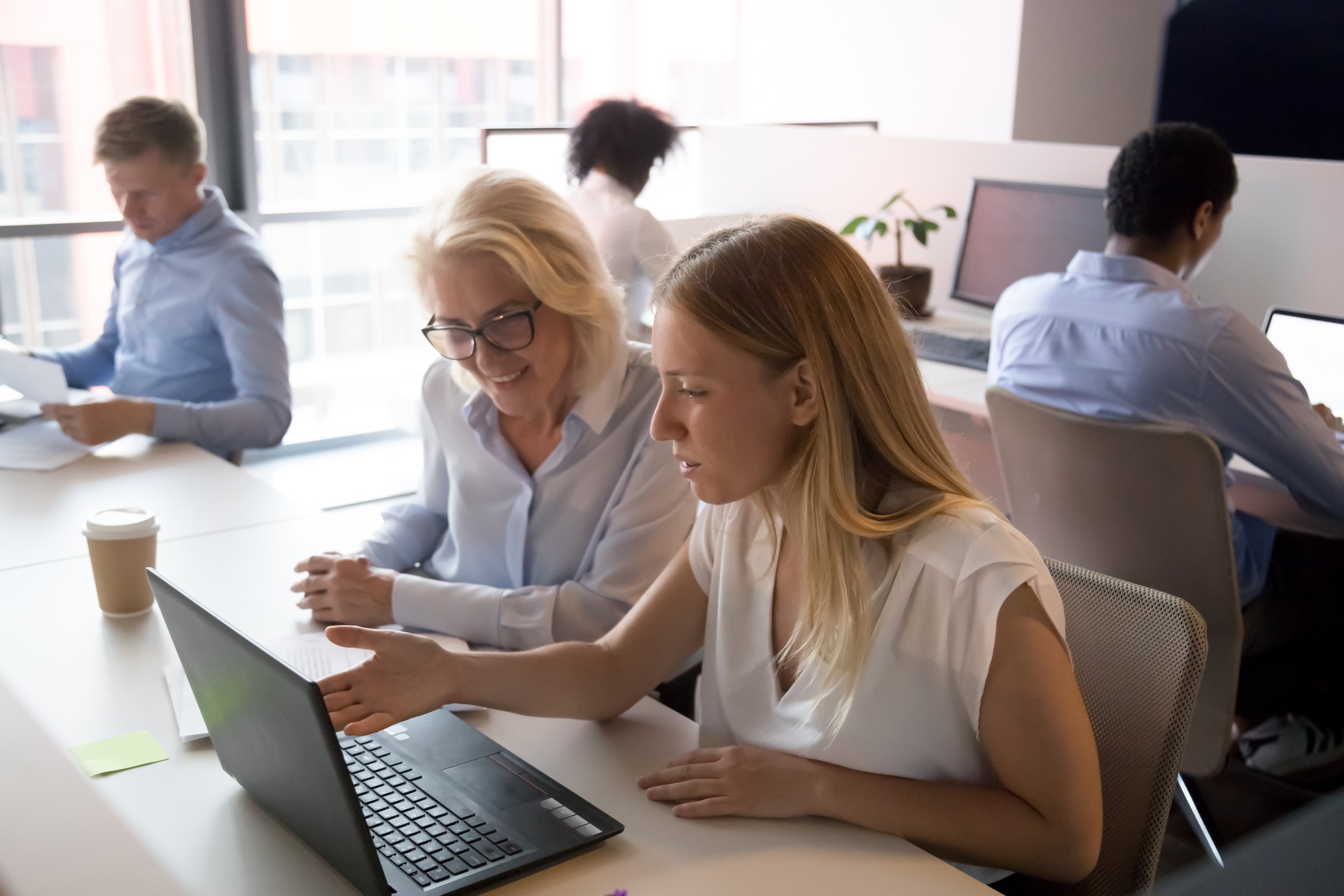 Young saleswoman manager mentor advisor consulting old female client or new employee pointing at laptop showing business presentation teaching instructing at meeting in modern corporate shared office