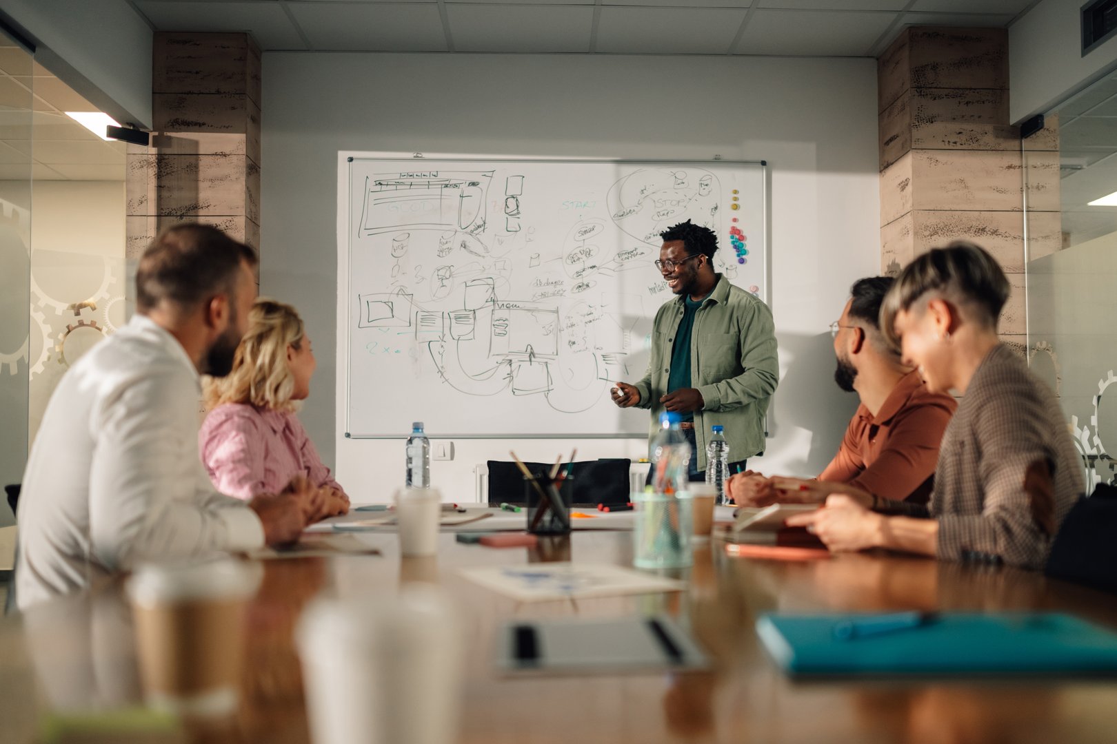 Diverse team of professionals is sitting at a large wooden table in a modern office, attentively listening to an african american businessman presenting a new project on a whiteboard