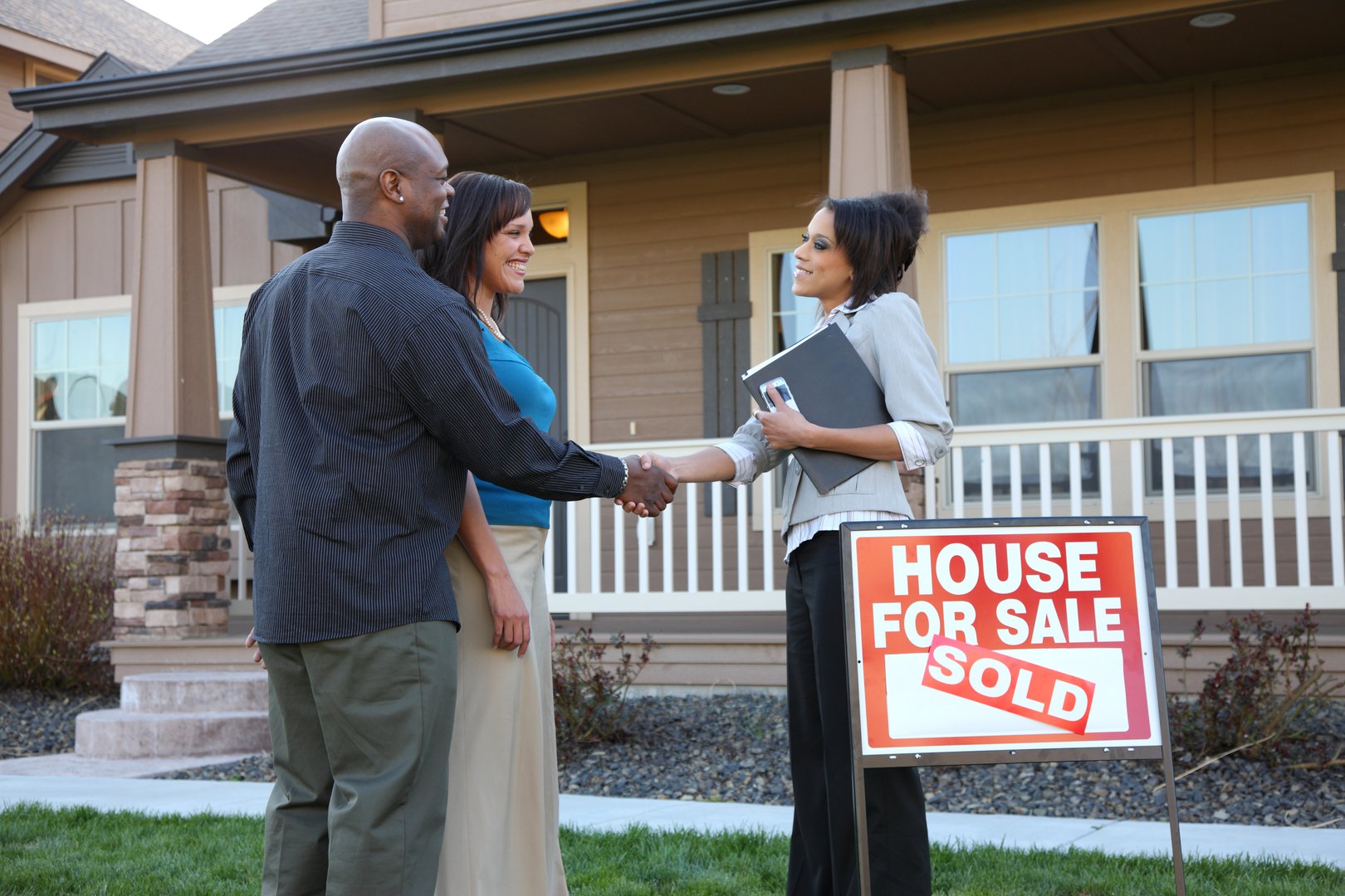 A couple shakes hands with a realtor in front of a house with a House for Sale sign marked Sold.
