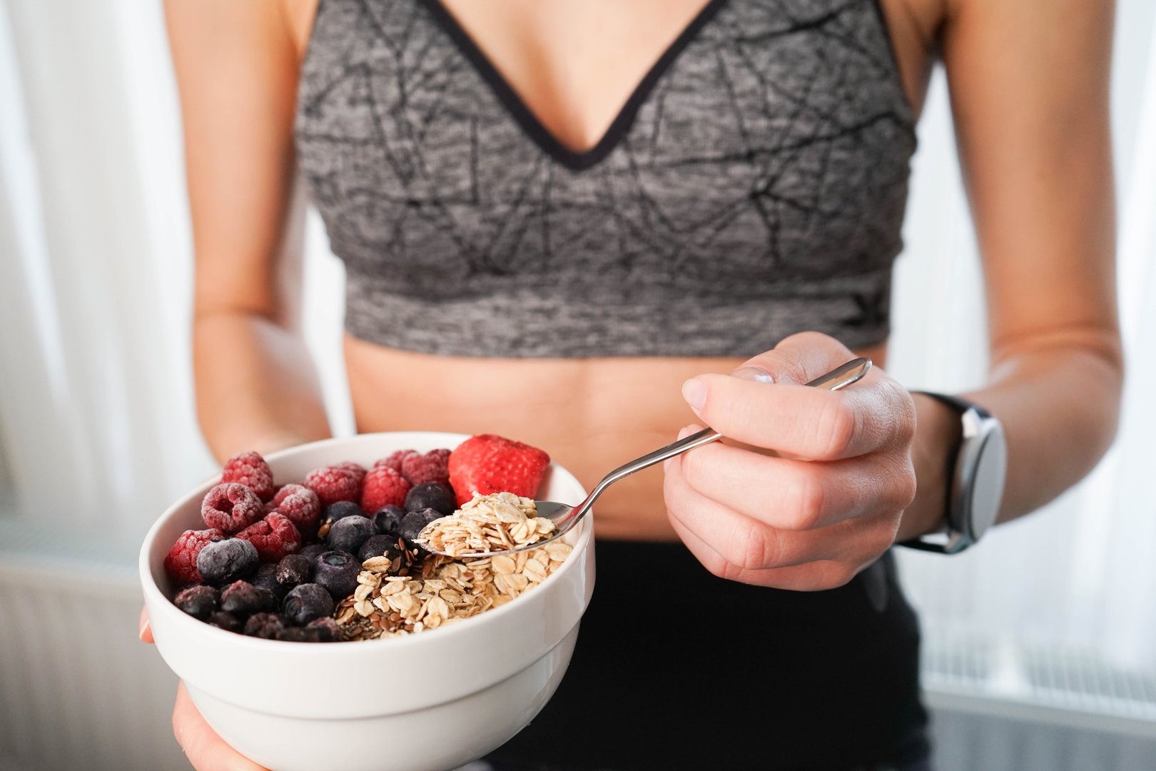A girl is eating oats with forest fruits at home after a fitness workout. She enjoys a healthy breakfast, sitting in sportswear, with fresh blueberries, raspberries, and strawberries making the oatmeal even more appealing.