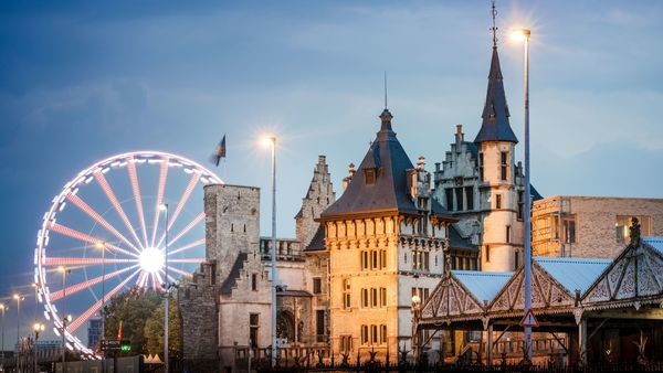 Medieval castle in Antwerp at dusk with a brightly lit Ferris wheel in the background, glowing under evening lights.