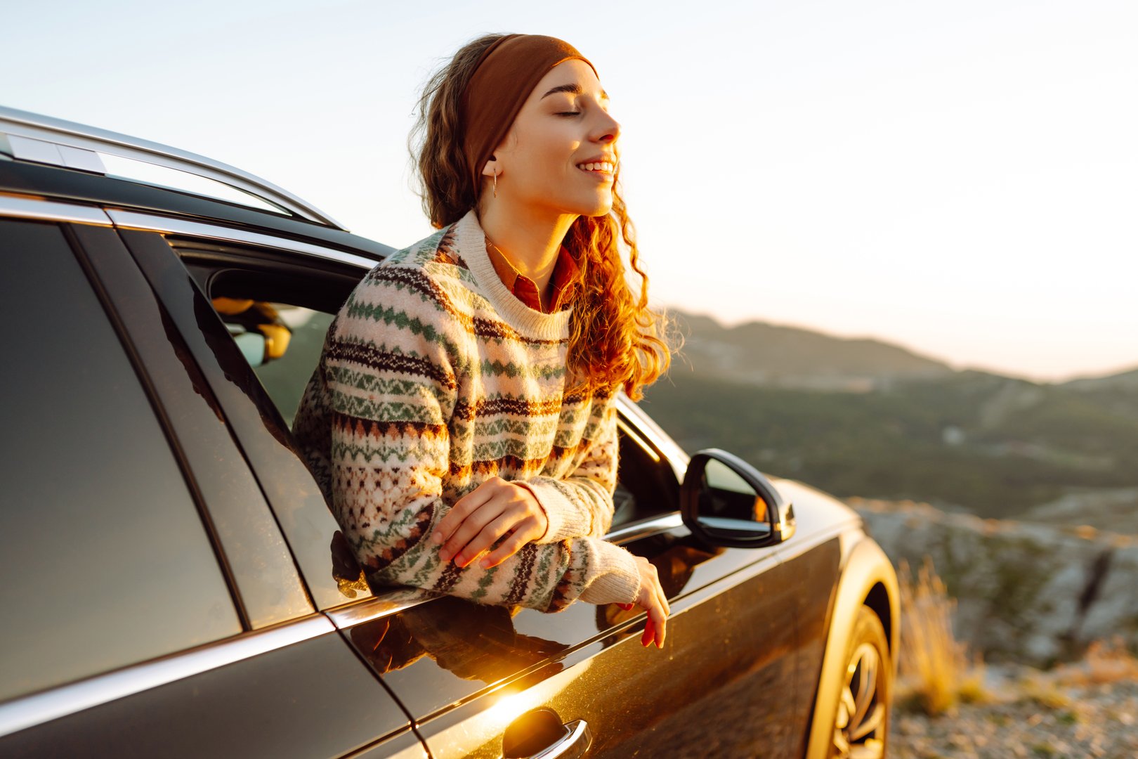 Happy woman standing near car and enjoys sunset. Summer trip. Lifestyle, travel, tourism, nature, active life.
