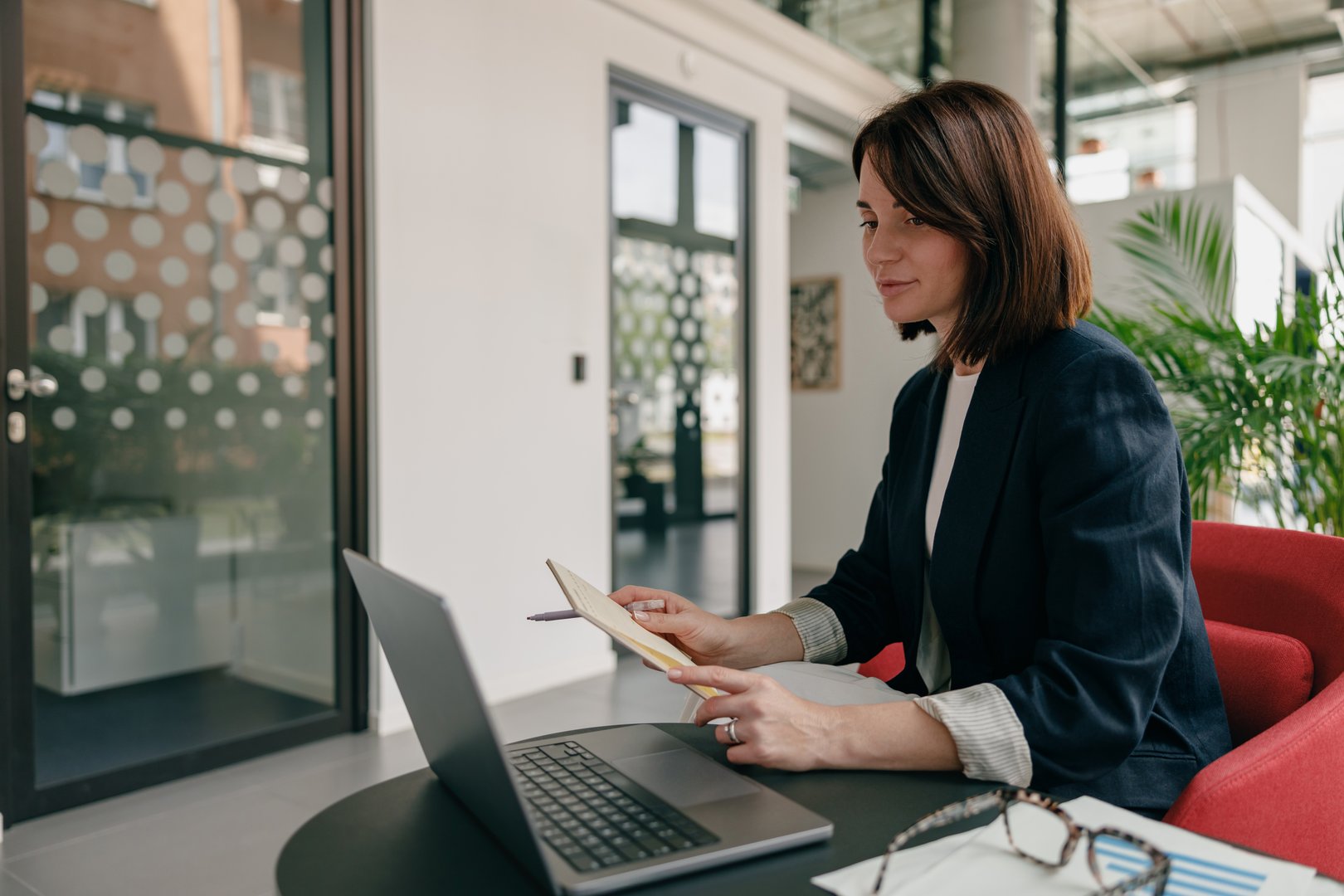 A businesswoman working remotely in a modern office setting, utilizing a laptop and notebook for her tasks.