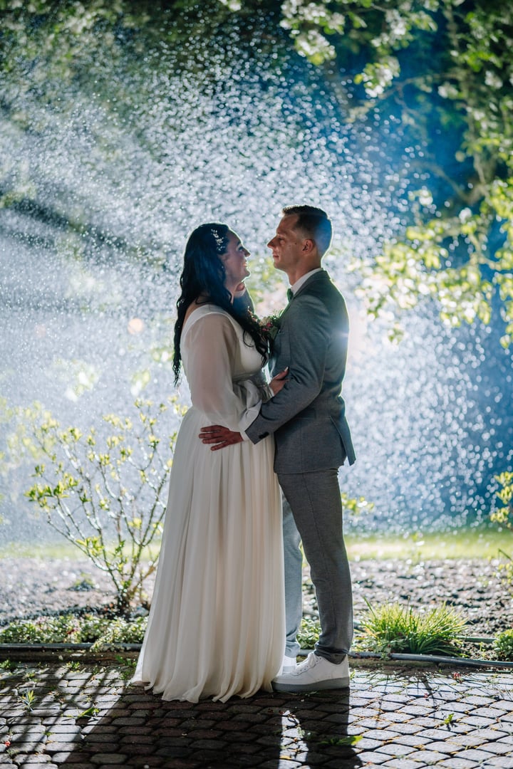 Couple in wedding attire share a tender moment surrounded by soft rain and illuminated by gentle lights, creating a romantic and enchanting atmosphere for their special day