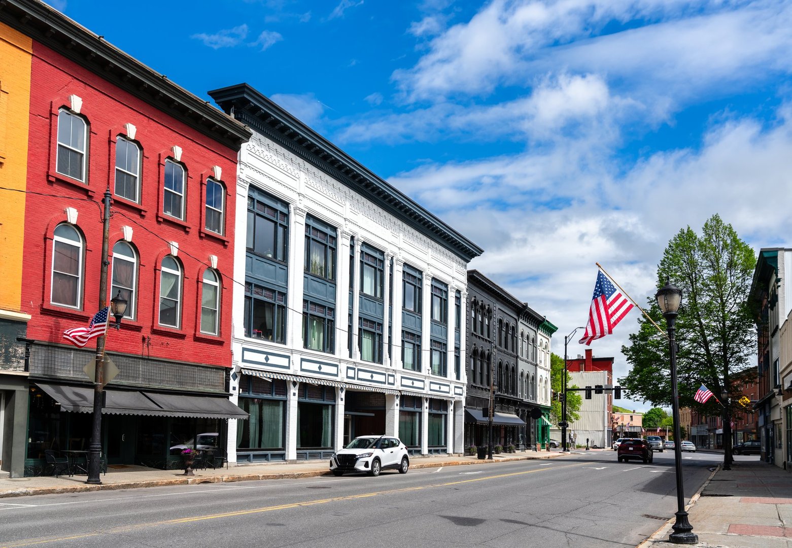 A row of red, white, and gray 19th-century storefronts lines Water Street in Augusta, Maine, United States, where American flags flutter beneath a bright spring sky.