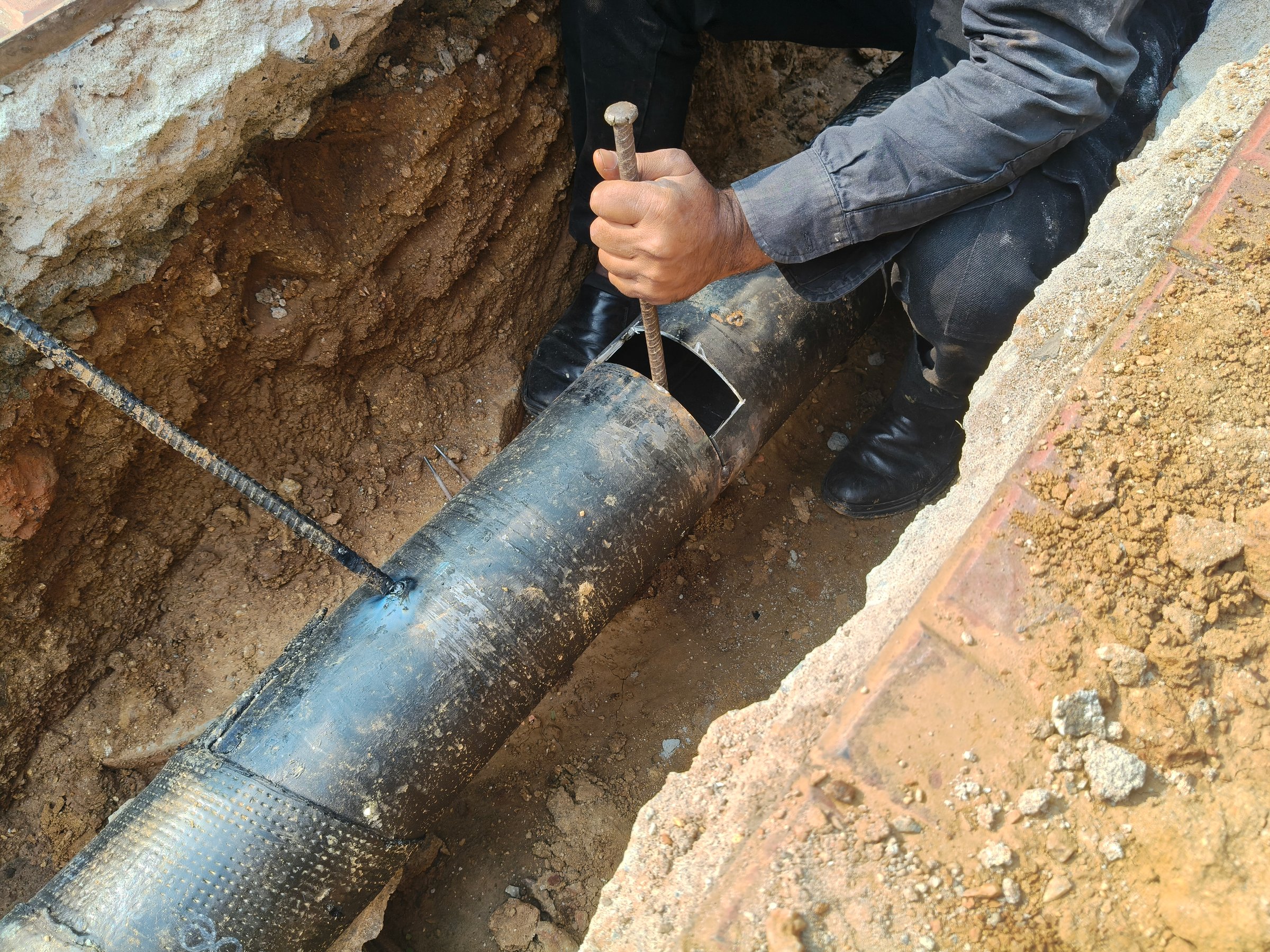A man is welding a thick iron pipe. The worker is welding a patch onto a large, dark colored pipe. All the welding equipment is present.