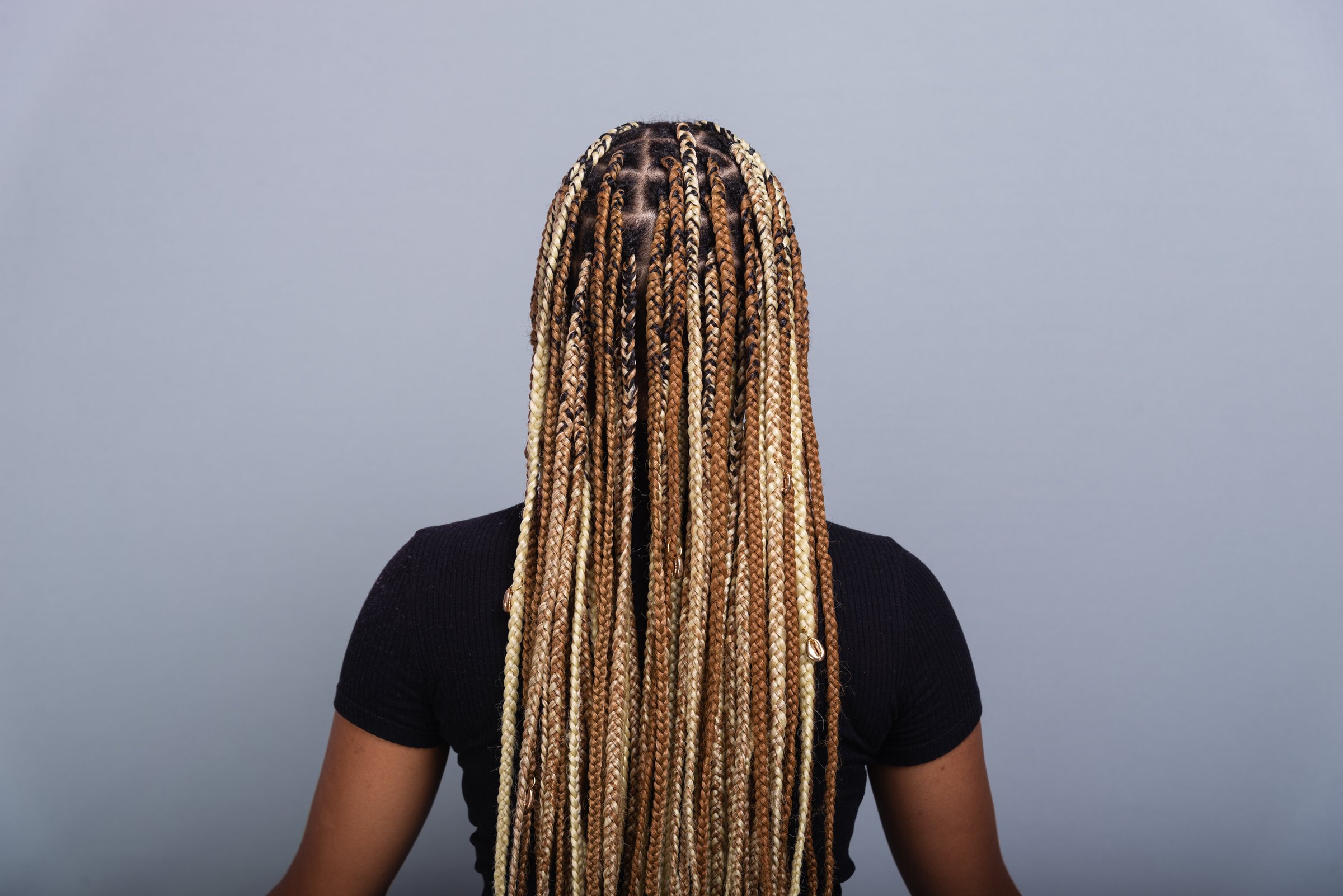 Brunette woman with braids in her hair, wearing jeans and a black blouse, sitting on the studio floor with her back to the camera. Isolated on a gray background.