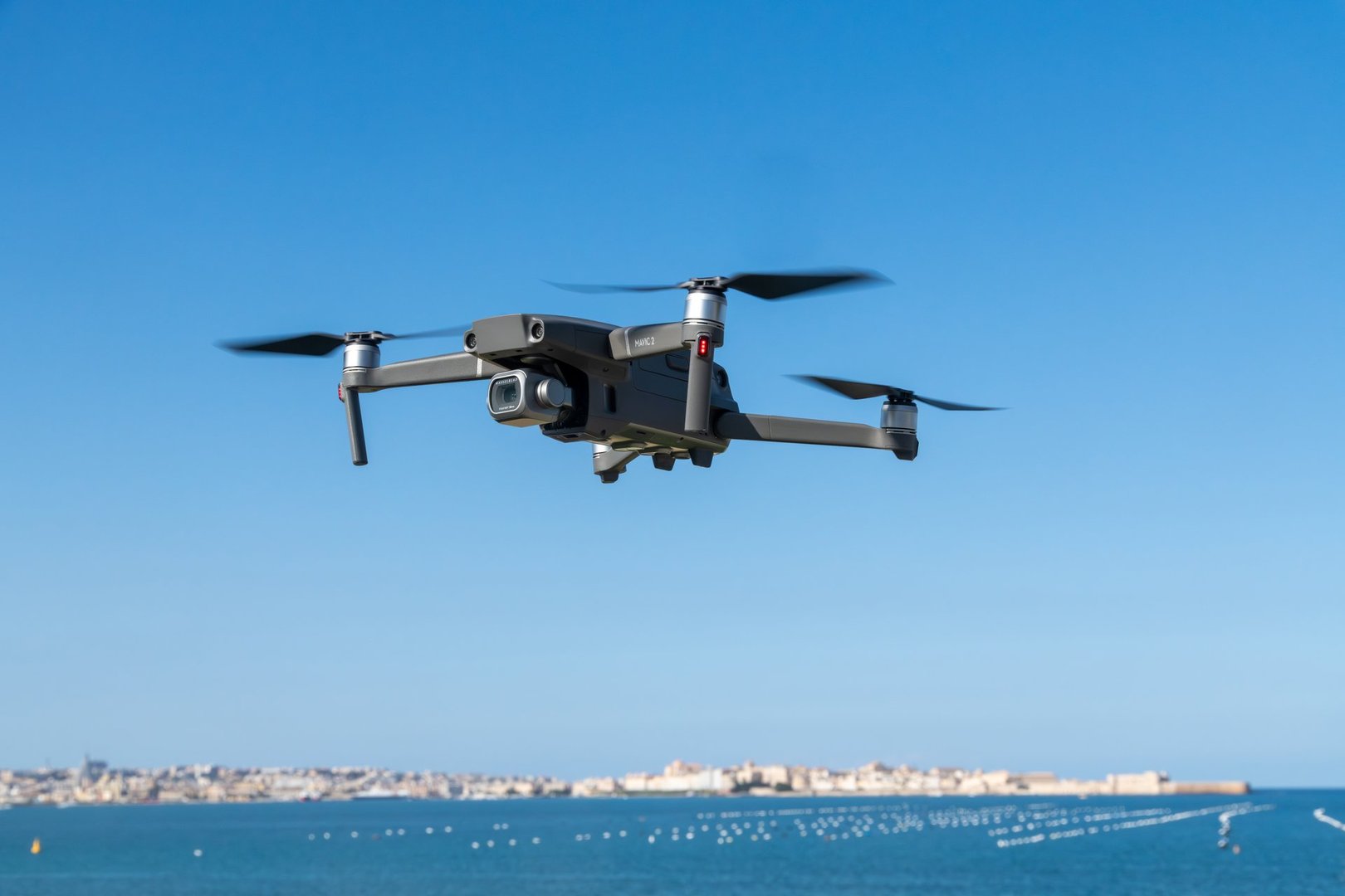 Syrakus, Sizilien - Italy - 04-10-2025: A drone hovers above the coast near Syracuse with spinning propellers, framed by calm blue sea and a distant skyline under clear sky.