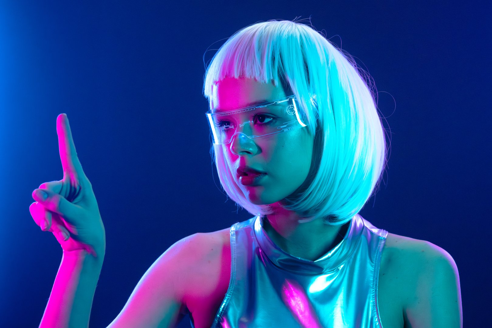 Stylish futuristic woman with white hair and transparent smart glasses, pointing upwards. She is illuminated by vibrant neon blue and pink lights, wearing a shiny silver outfit in a cyberpunk theme.