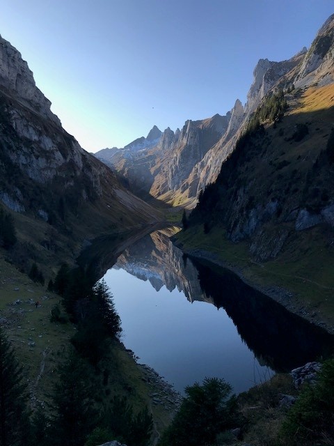 Scenic view of a mountain valley with a calm reflective lake, surrounded by steep rugged cliffs and forested slopes.