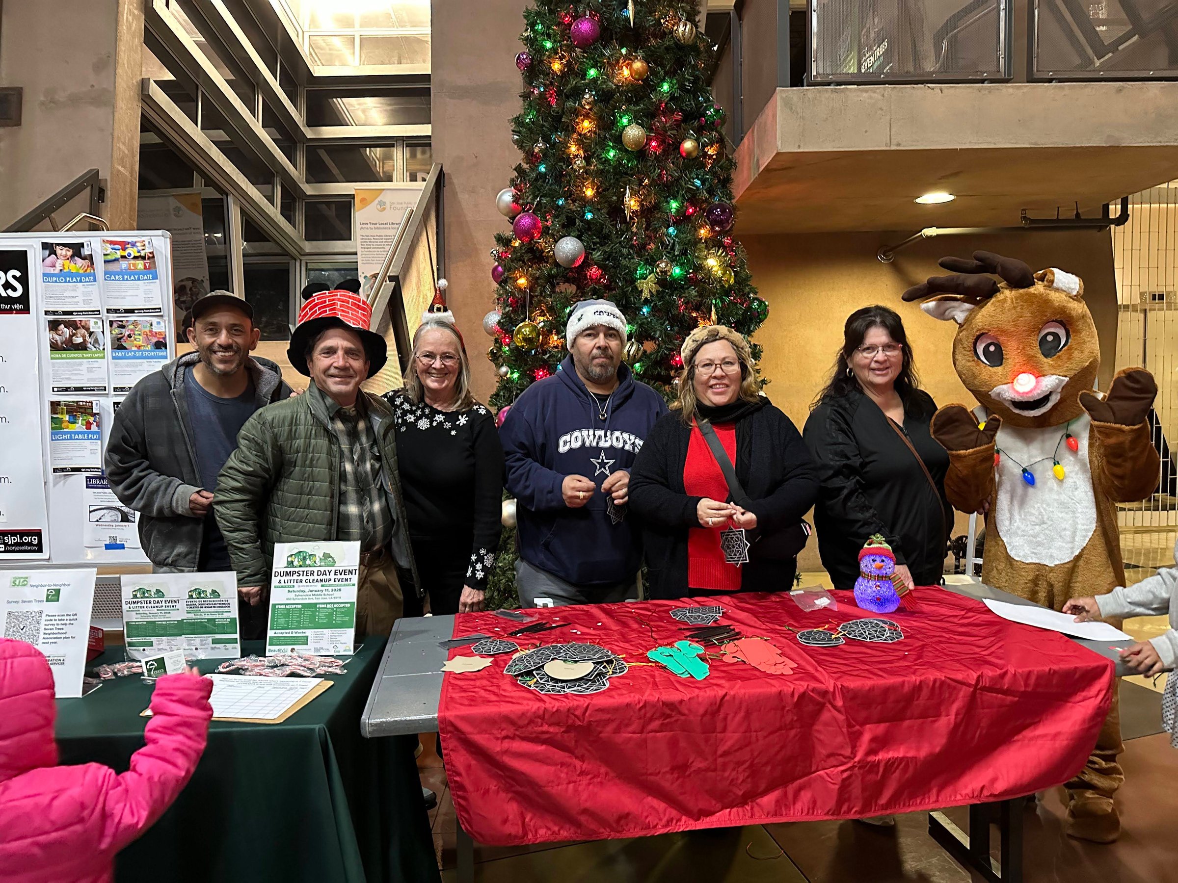 People at a festive event table with holiday decor, a Christmas tree, and a person in a reindeer costume.