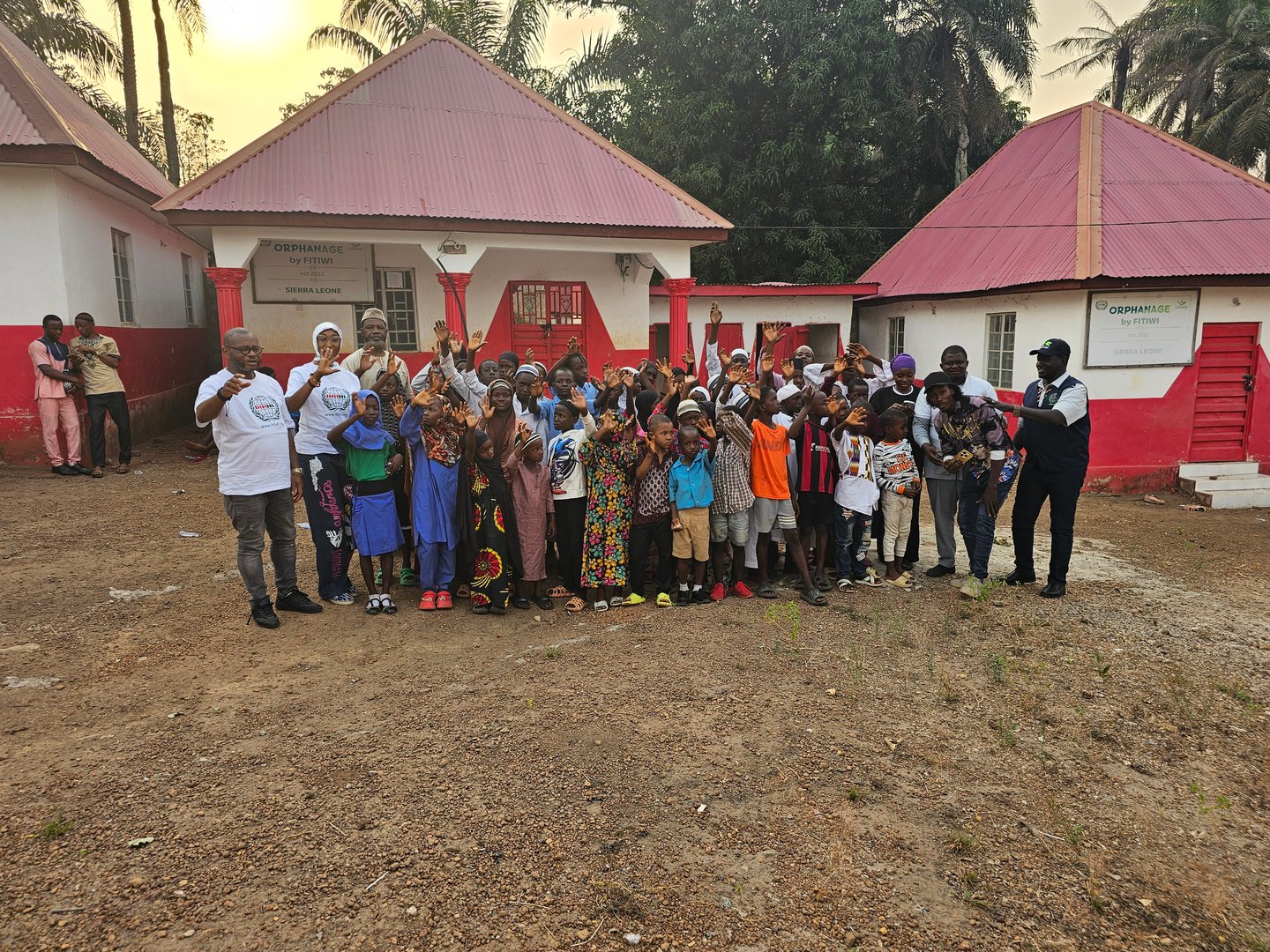 Group of children and adults posing in front of a building with red roofs, surrounded by trees, during the day.