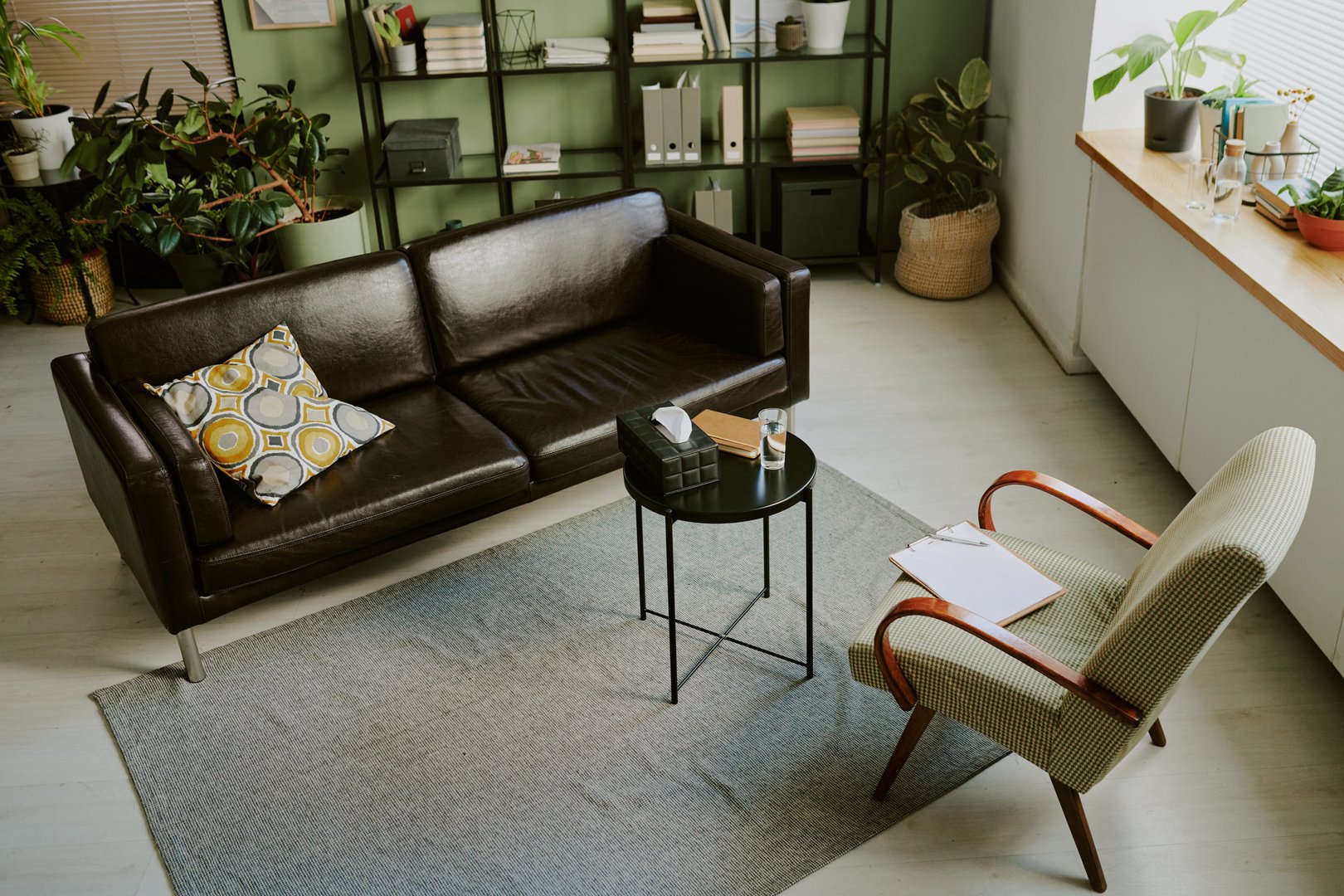 Cozy office lounge area featuring a brown leather couch and an armchair with a side table, indoor plants, bookshelves, and natural light coming through window