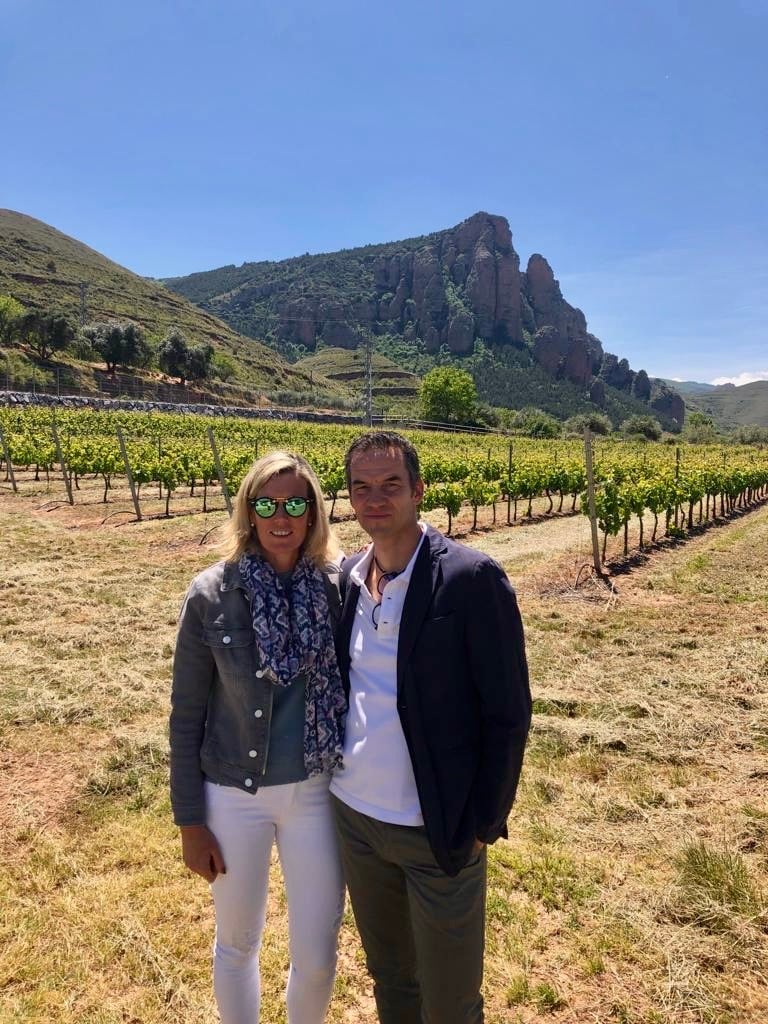 A couple standing in a vineyard with a rocky hill in the background on a sunny day.