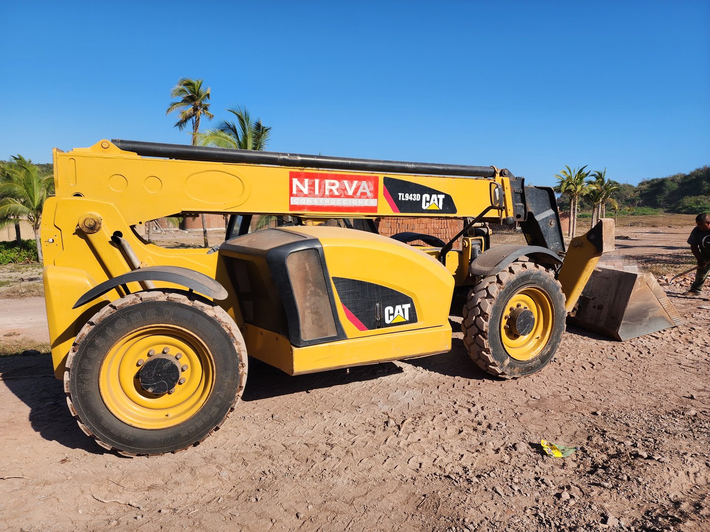 Yellow CAT telehandler on a dirt road with palm trees in the background, sunny day.
