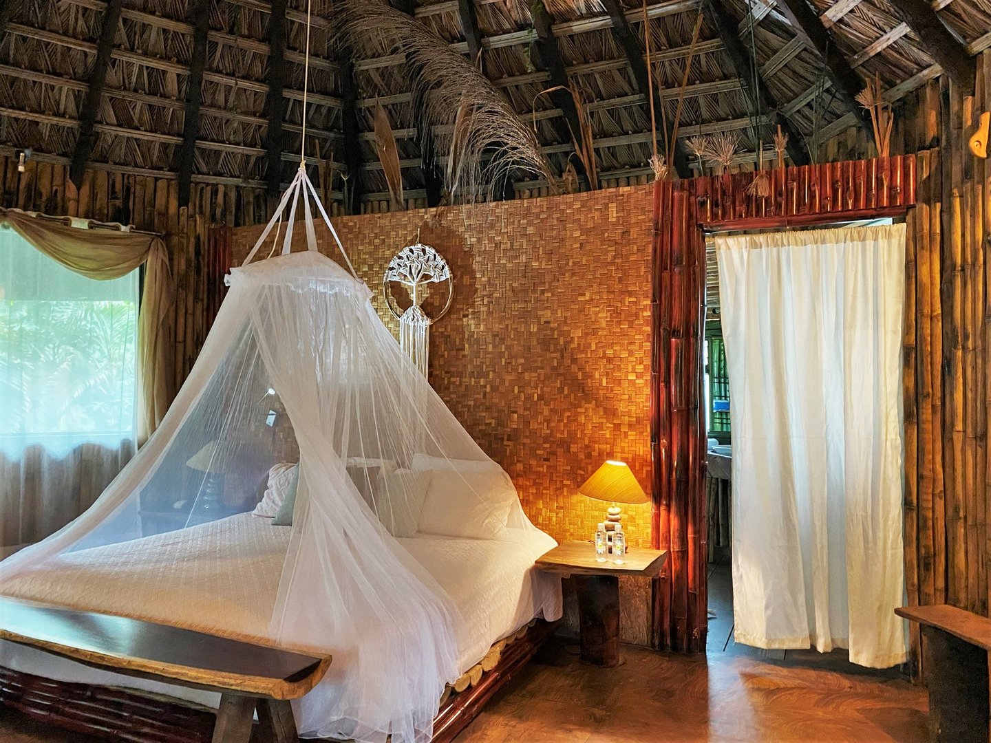A cozy bedroom with a mosquito-netted bed, wooden walls, and a warm lamp next to it, creating a rustic atmosphere.