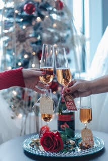 Two hands clinking champagne glasses in front of a decorated Christmas tree, with a table holding a bottle and rose.