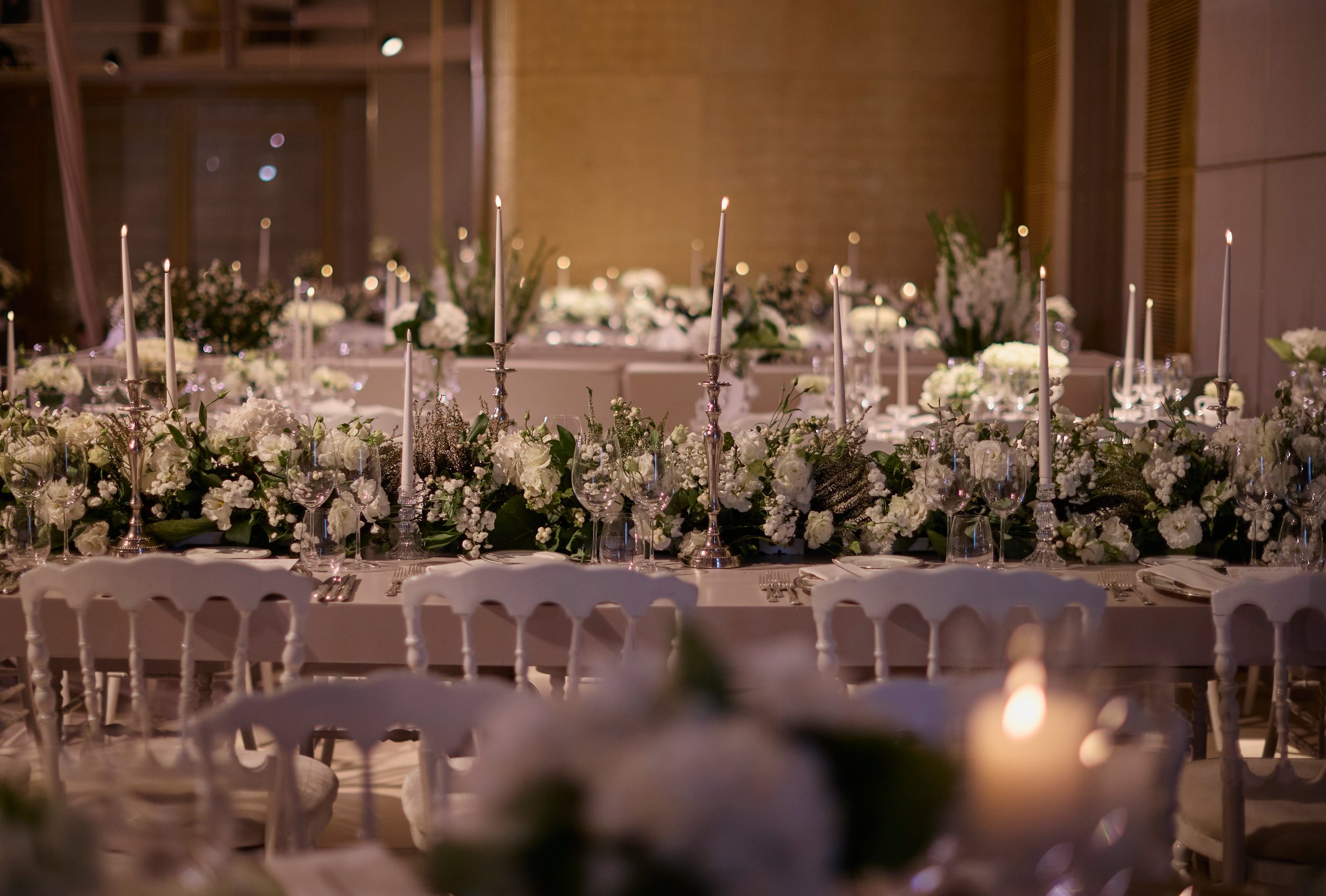 A beautifully set wedding dinner table featuring tall, slim candles burning in elegant silver candleholders, complemented by white roses arranged in crystal vases. The combination of soft candlelight and delicate flowers creates a sophisticated and romantic atmosphere, perfect for special occasions, weddings, and elegant event decor.