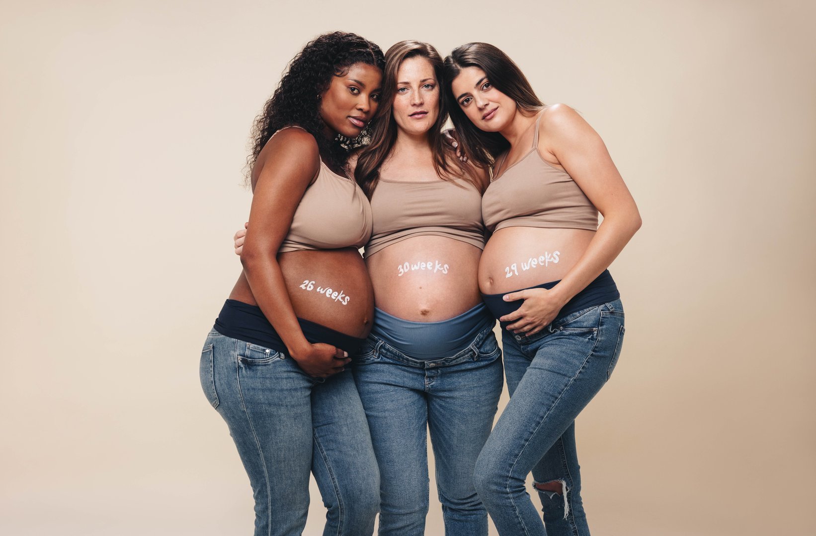 Young, diverse pregnant women in the third trimester stand together in a studio. They wear jeans and bras, showcasing their baby bumps. Hugging and looking at the camera, they represent a supportive multicultural maternity group.
