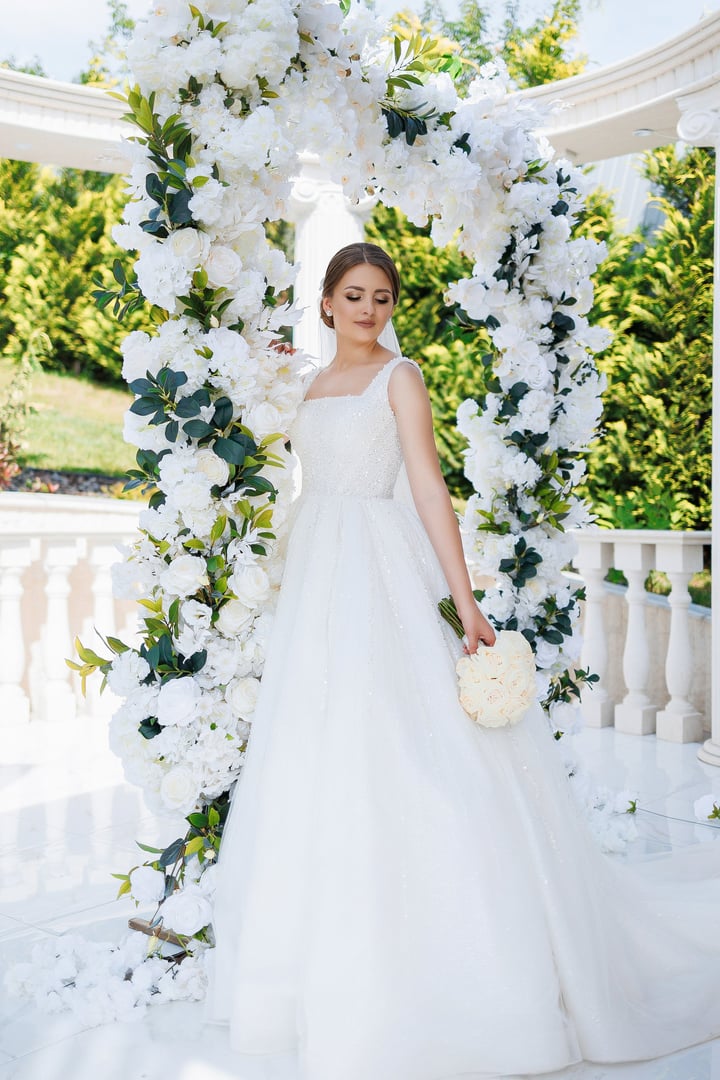 A bride is standing in front of a white archway, holding a bouquet of flowers. The archway is decorated with white flowers, creating a romantic and elegant atmosphere