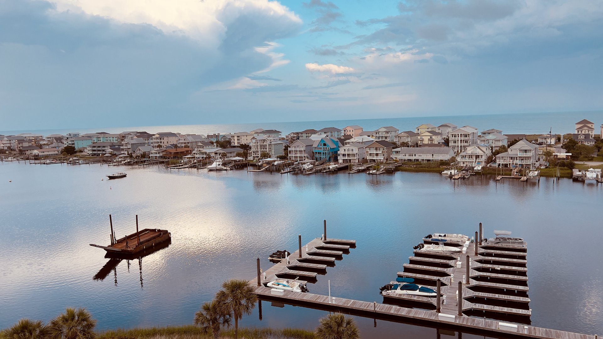 Barge on a North Carolina coastal waterway with view of the Atlantic Ocean. The deck barge has a mirrored reflection in the water. There are palm trees in the foreground and other boats nearby. The water is calm, and the vast sky has some clouds. This is a natural scene, no filters or color edits were used.