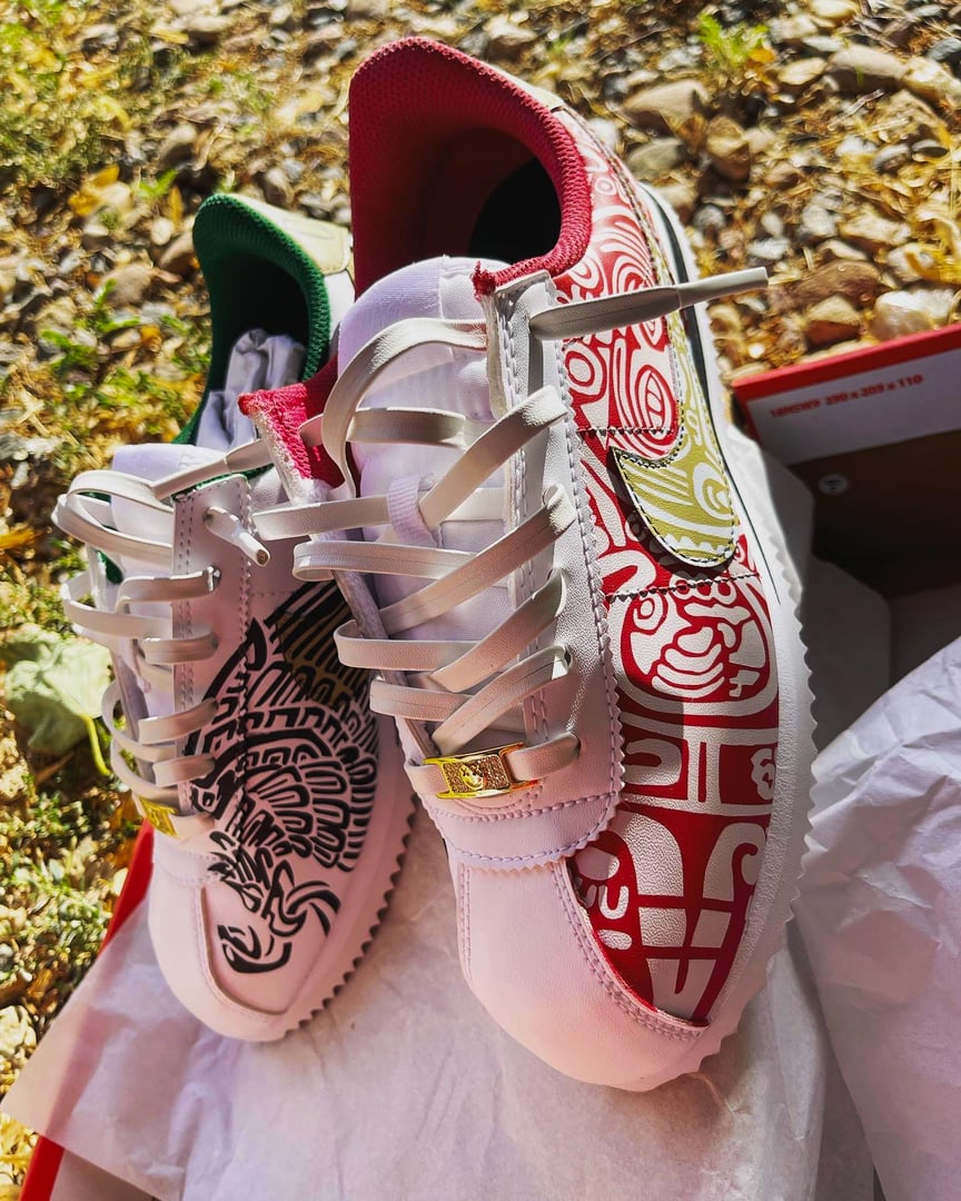 Two colorful sneakers with intricate designs, featuring red and black patterns, displayed outdoors on paper and grass.