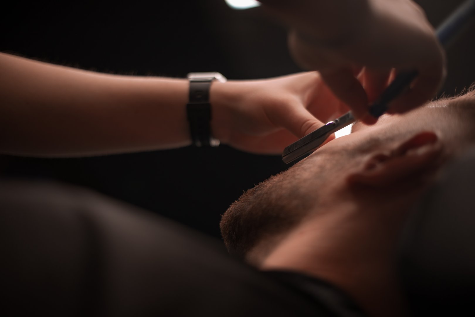 Close-up view of a barber shaving a client's cheek with a straight razor. The barber's hands and the client's trimmed beard are in focus, capturing the precision and care of the grooming process.
