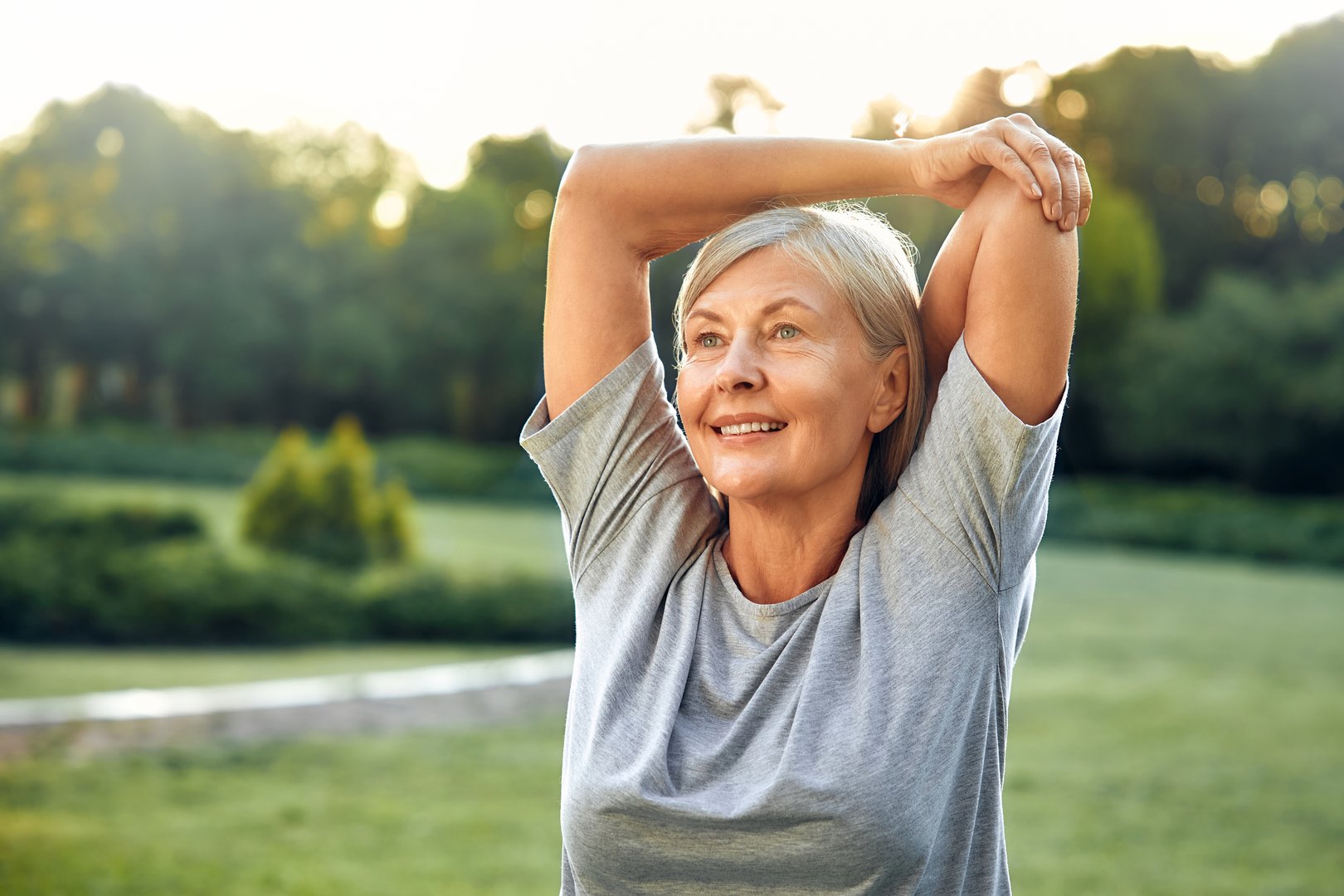 A joyful senior woman is gracefully stretching her arms outdoors in a lush, expansive green park, fully embracing a life of wellness, vitality, and the beauty of nature around her