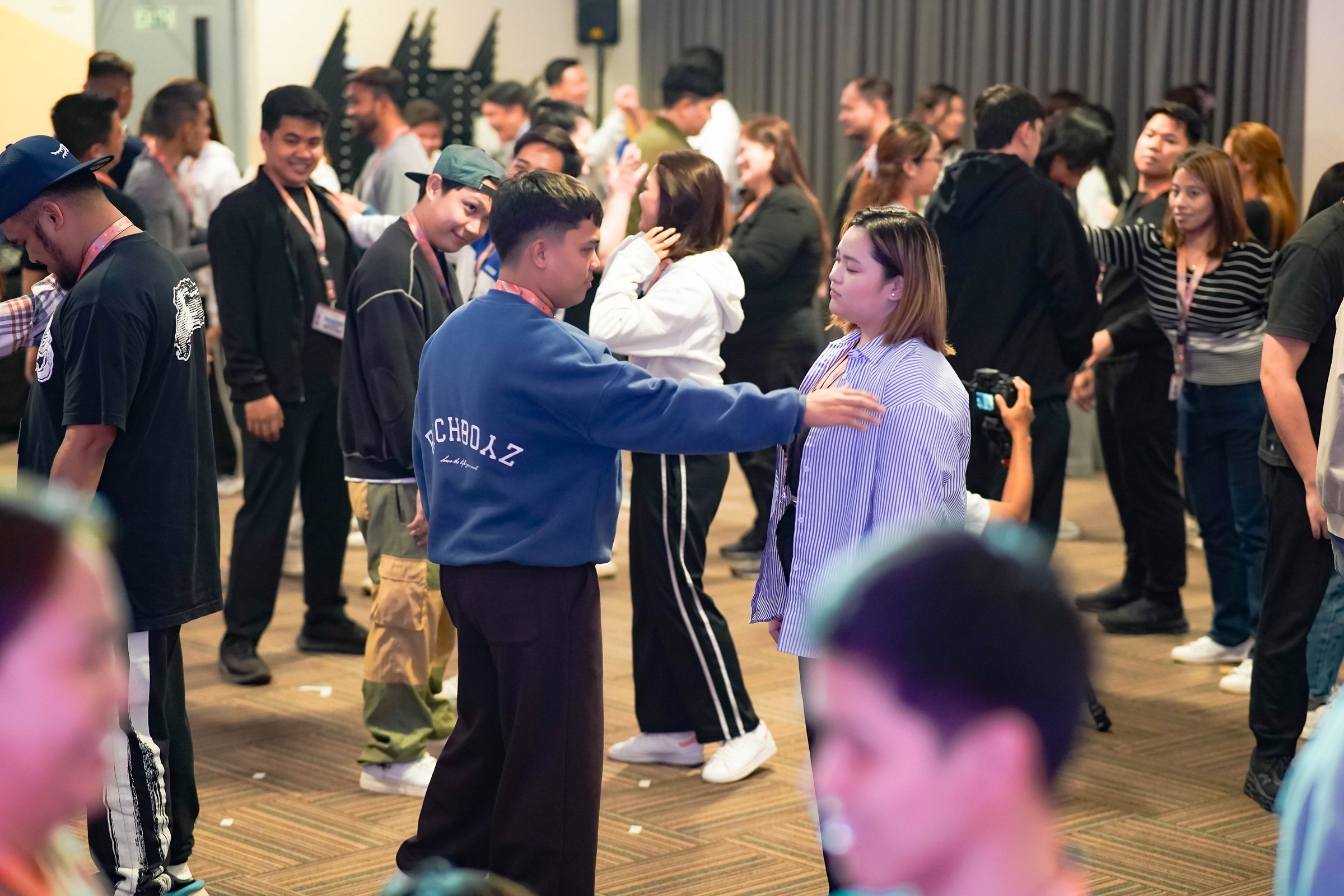 People engaging in a group activity in a large room, talking and interacting, with a mix of casual outfits.