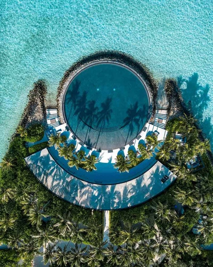 Aerial view of a circular pool surrounded by palm trees and clear turquoise water, creating a tropical paradise setting.