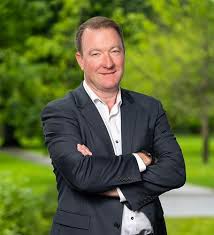 Man in a dark suit with arms crossed stands outdoors against a lush green park background.