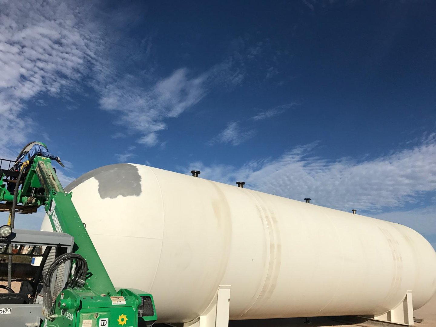 A large white cylindrical storage tank under a blue sky with thin clouds, partially obscured by green construction equipment.