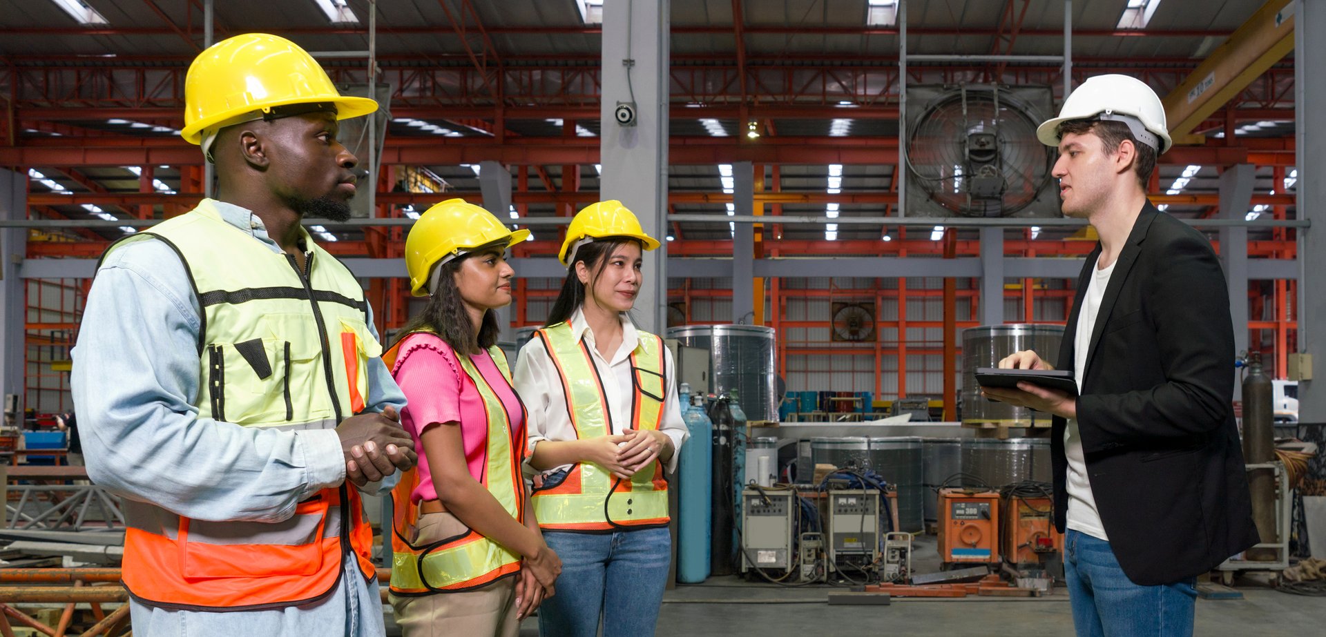 Group of engineer in safety vest meeting with young manager in black suit in an industrial or factory setting. Everyone wearing protective hardhat. Atmosphere in manufactory with large machinery.