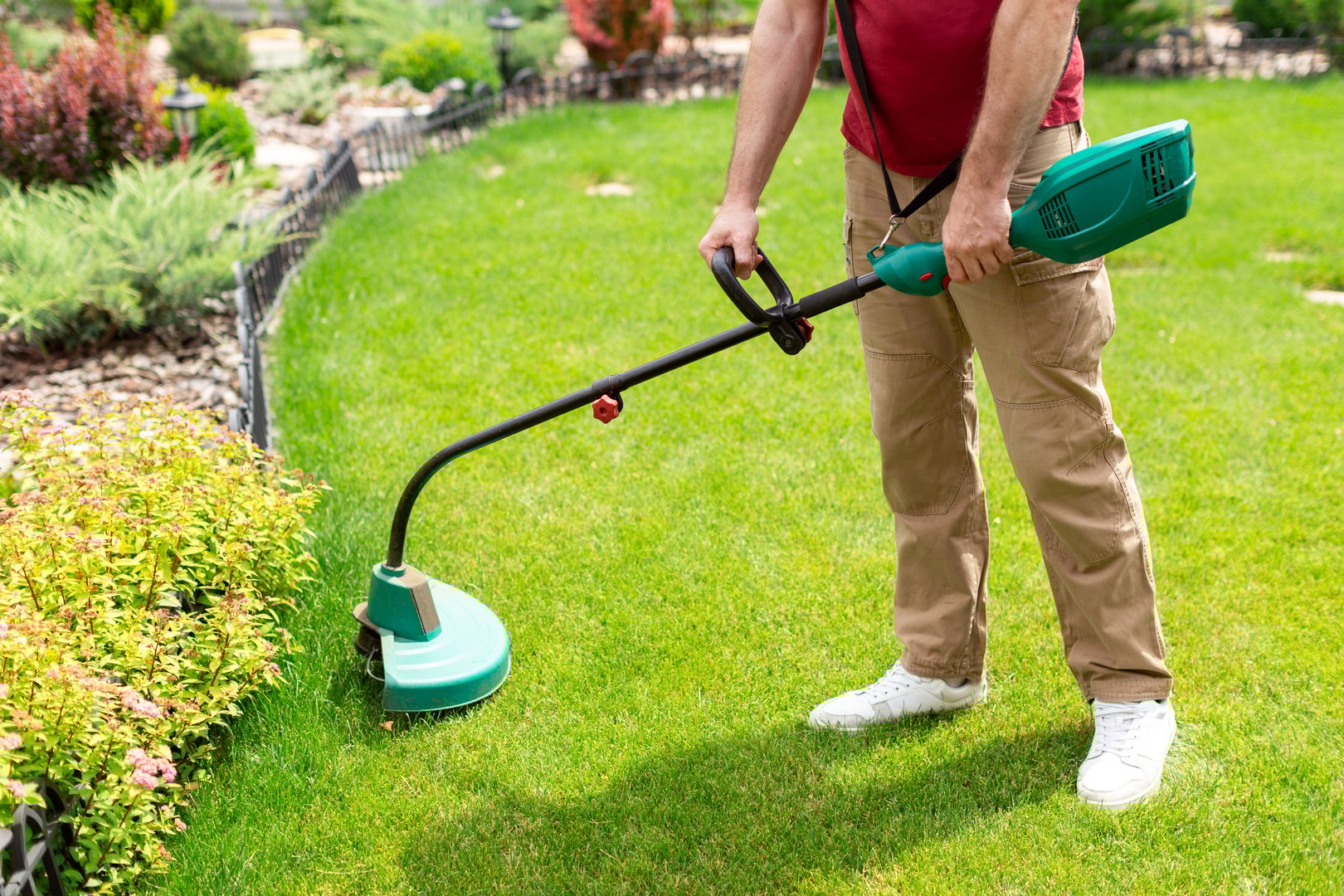 Unrecognizable man gardener using electric weed trimmer to trim grass in his garden. Lawn care in backyard